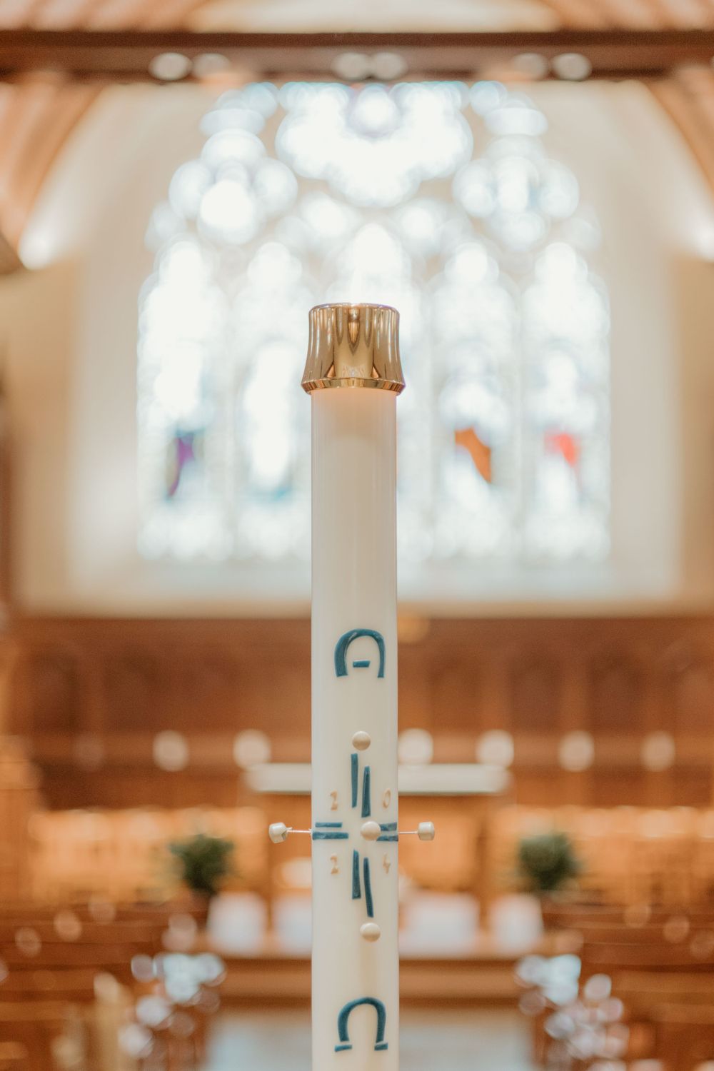 White candle with religious symbols stands in front of stained glass window inside church sanctuary.