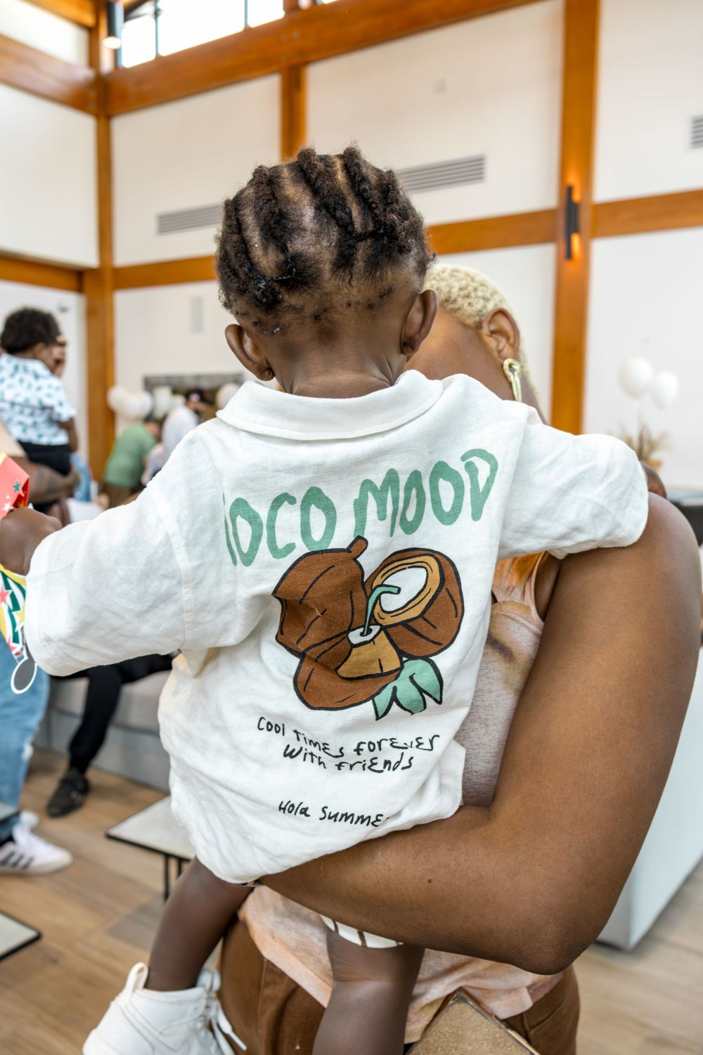 Child wearing a white t-shirt with 'Coco Mood' graphic print being held at an indoor event.