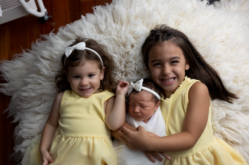 Three children wearing yellow and white dresses pose together on a plush white fur rug - straight out of camera