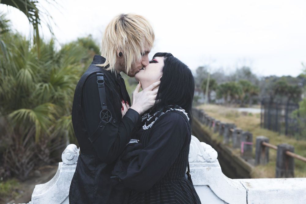 Romantic gothic couple in black clothing embracing near palm trees.