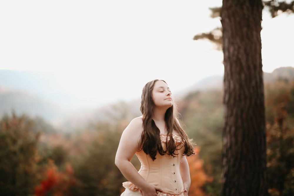 Woman in vintage corset dress poses against autumn mountain landscape in warm sunset light.