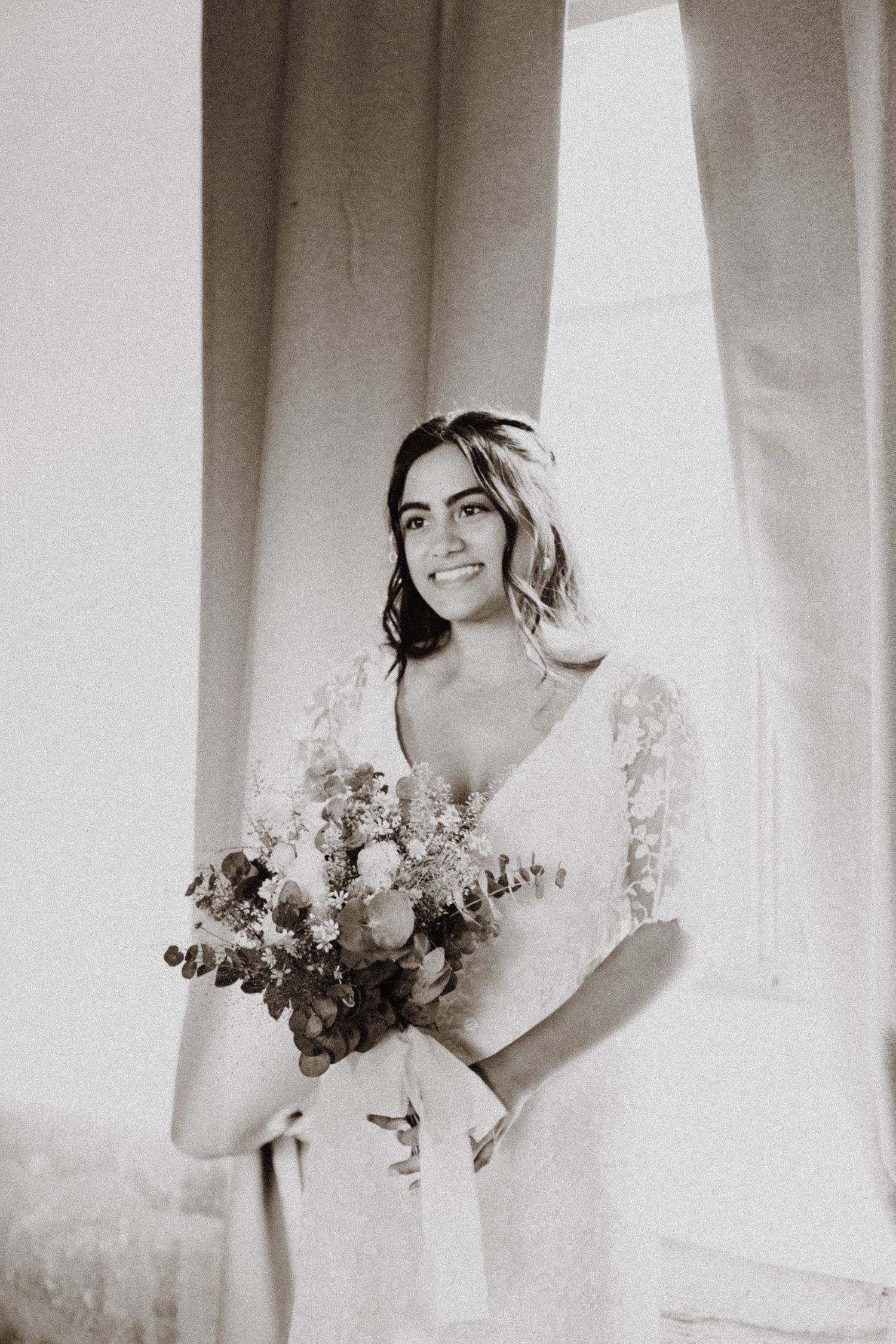 Black and white portrait of bride holding bouquet against light-filled window.