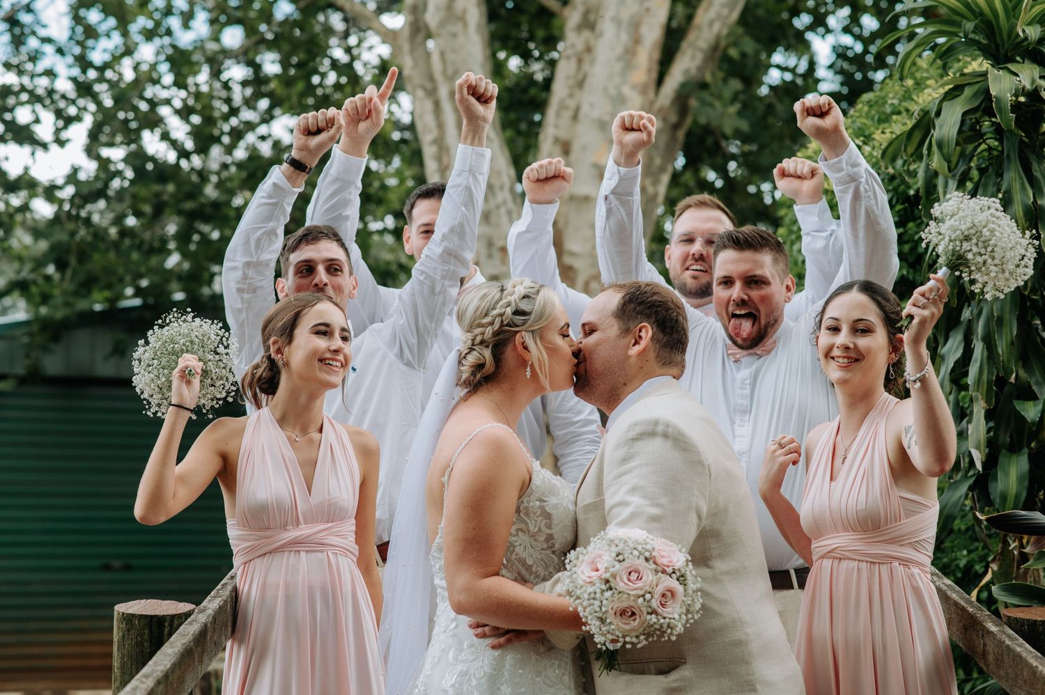 Wedding party celebrating on wooden bridge with raised arms as newlyweds share a kiss.
