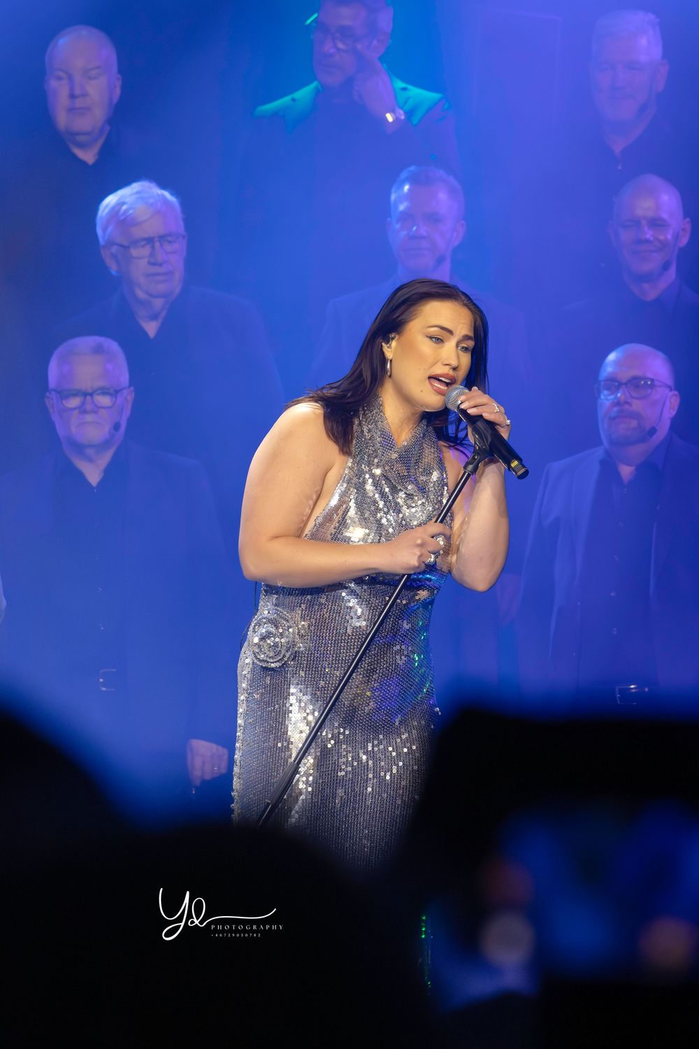 A singer in a sparkly silver dress performs on stage with a choir visible in blue lighting behind her.