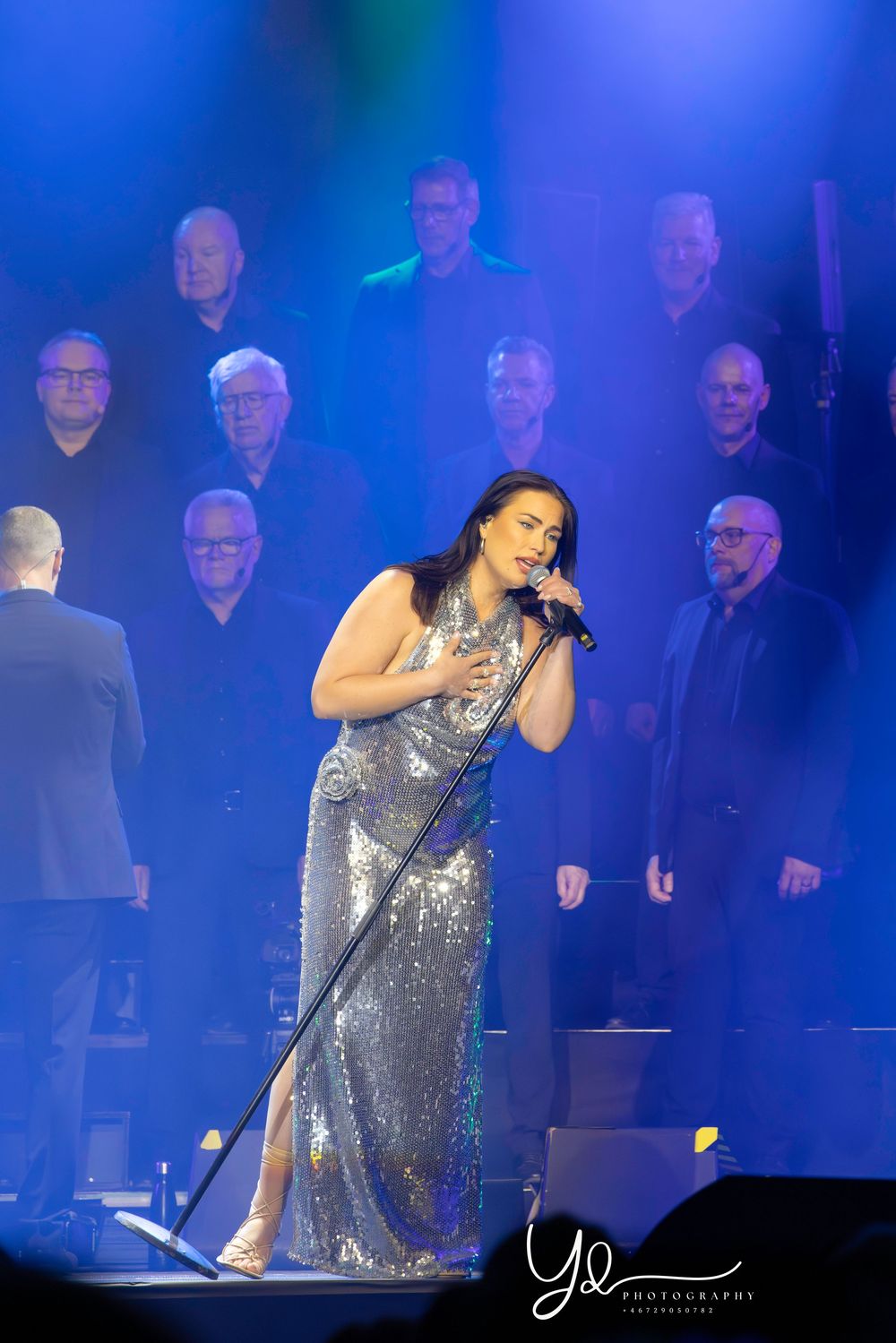 Singer in a sparkling silver dress performs passionately on stage with dark silhouettes and blue lighting in the background.