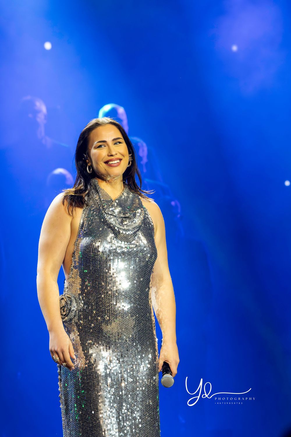 A performer in a sparkling silver sequin dress sings on stage against a vibrant blue backdrop.