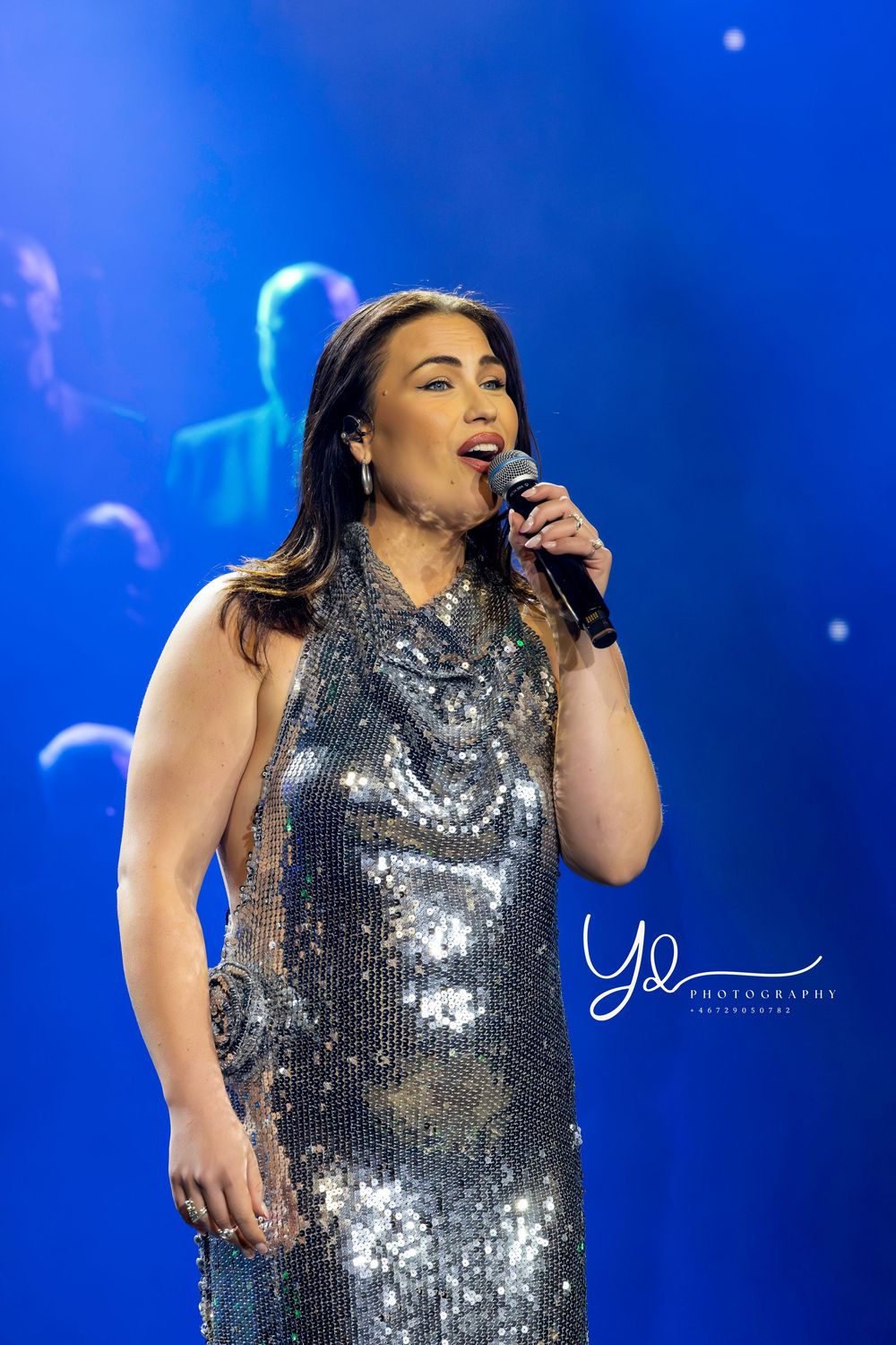 A singer wearing a glittering silver gown performs passionately against a deep blue stage backdrop.