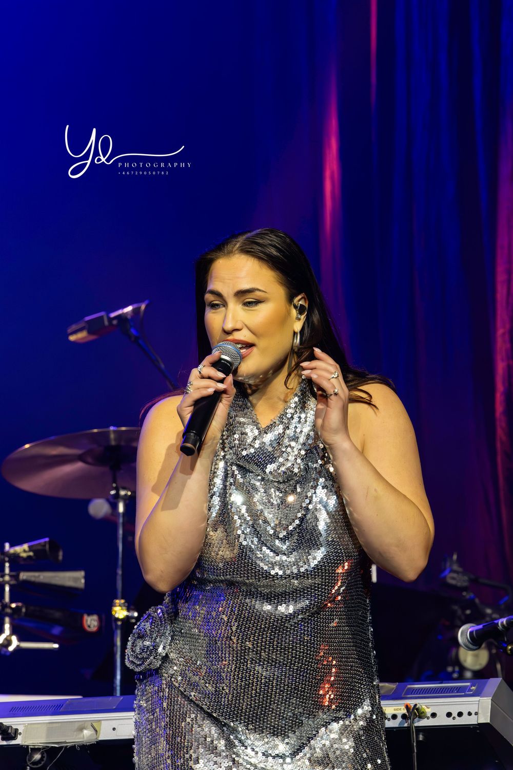 Singer performs in a sparkly silver dress on stage with microphone and instruments in the background against blue lighting.