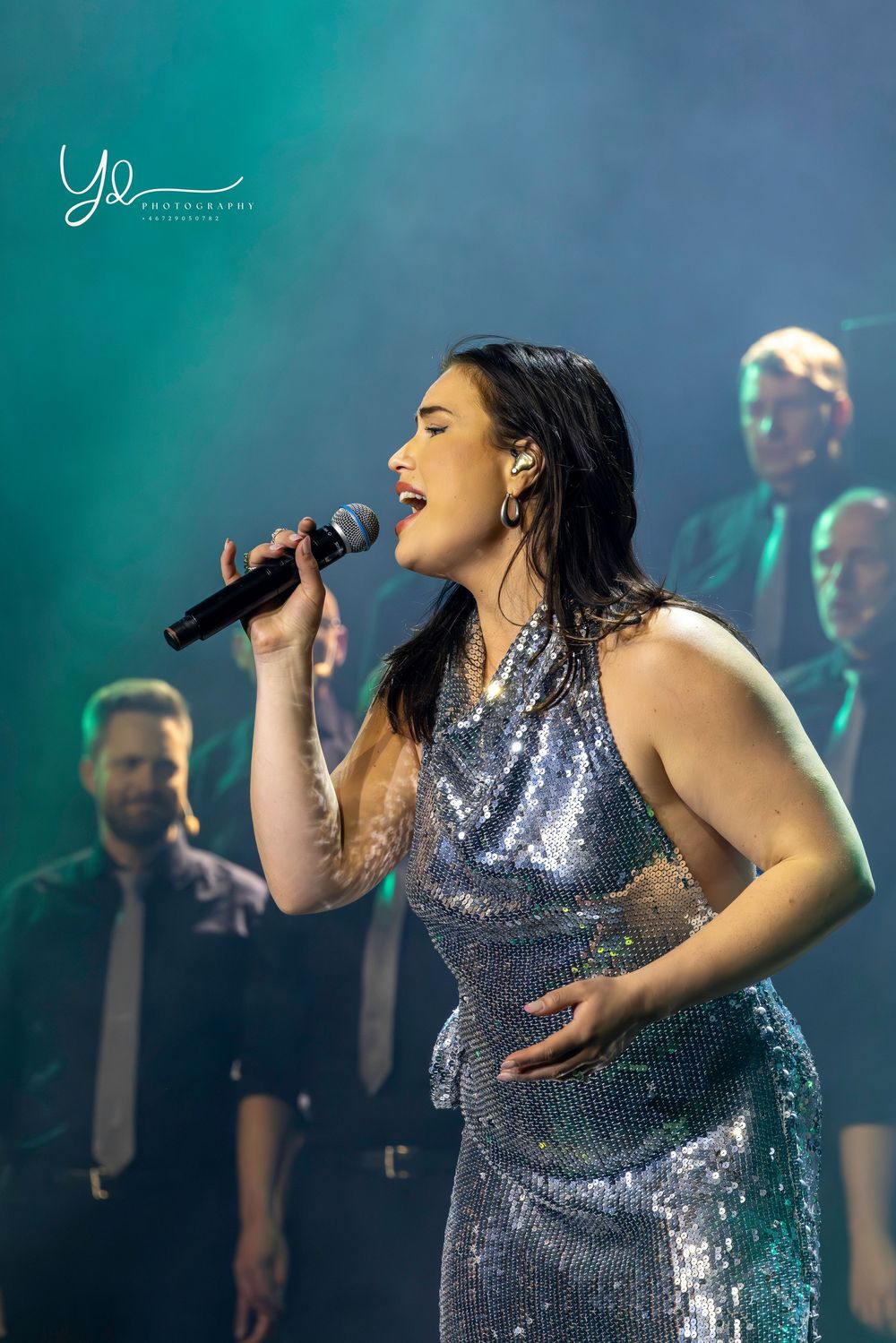 A performer in a sparkling silver dress sings passionately into a microphone on stage with backup singers visible in dark lighting.