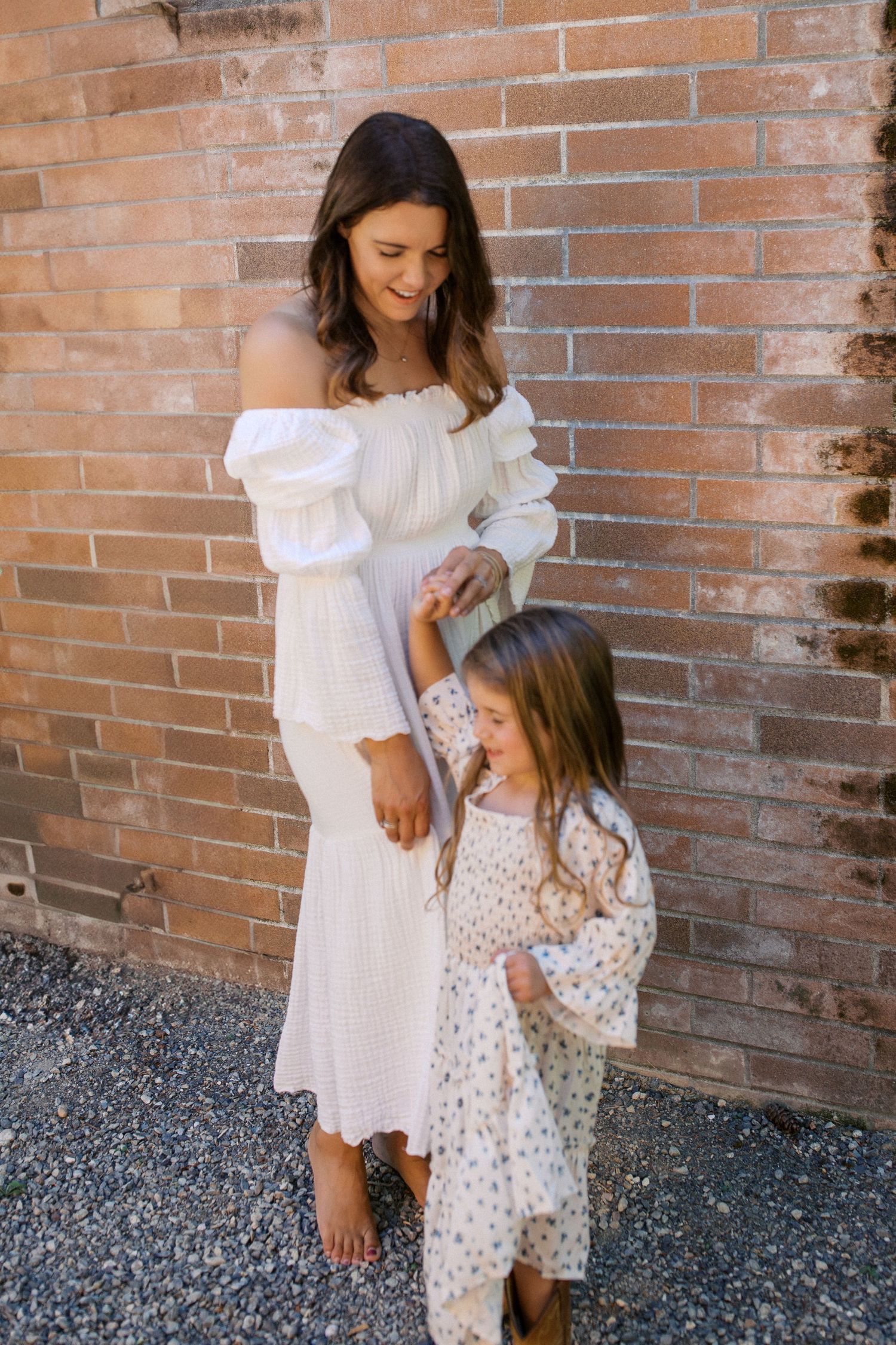 A mother in a white off-shoulder dress holds hands with her young daughter in a floral dress by a brick wall.