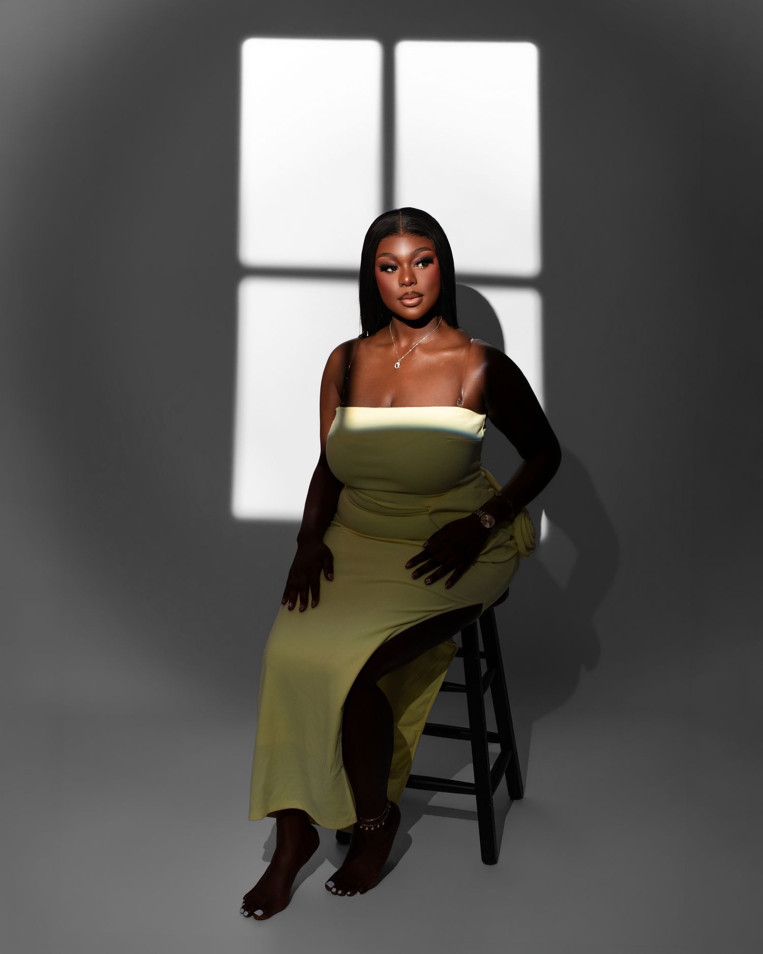 A woman in an olive green dress poses elegantly on a stool against a dramatic window-lit backdrop with strong shadows.