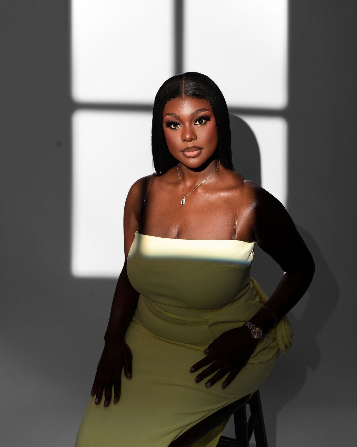 A fashion portrait in dramatic lighting shows a model in a yellow strapless dress and long black gloves against a shadowy backdrop.