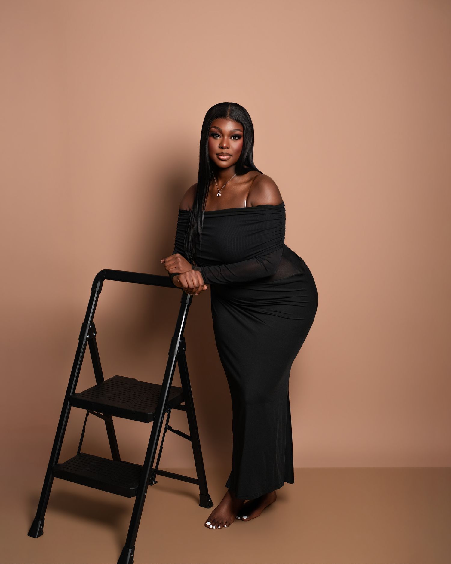 Fashion model in black dress poses elegantly with folding chair against beige backdrop in studio photoshoot series.