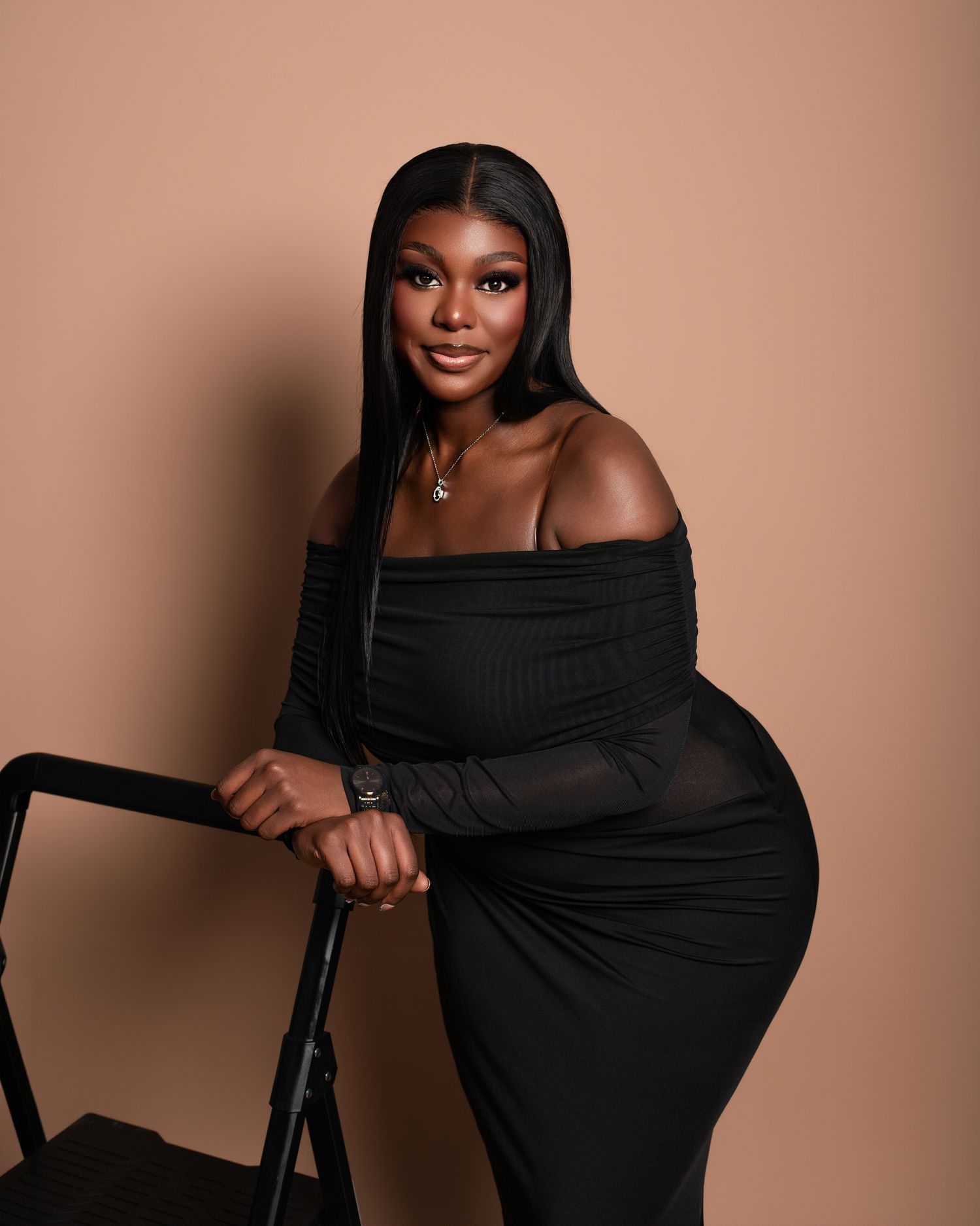 Studio portrait of a person in a black off-shoulder dress posing elegantly against a warm brown background.
