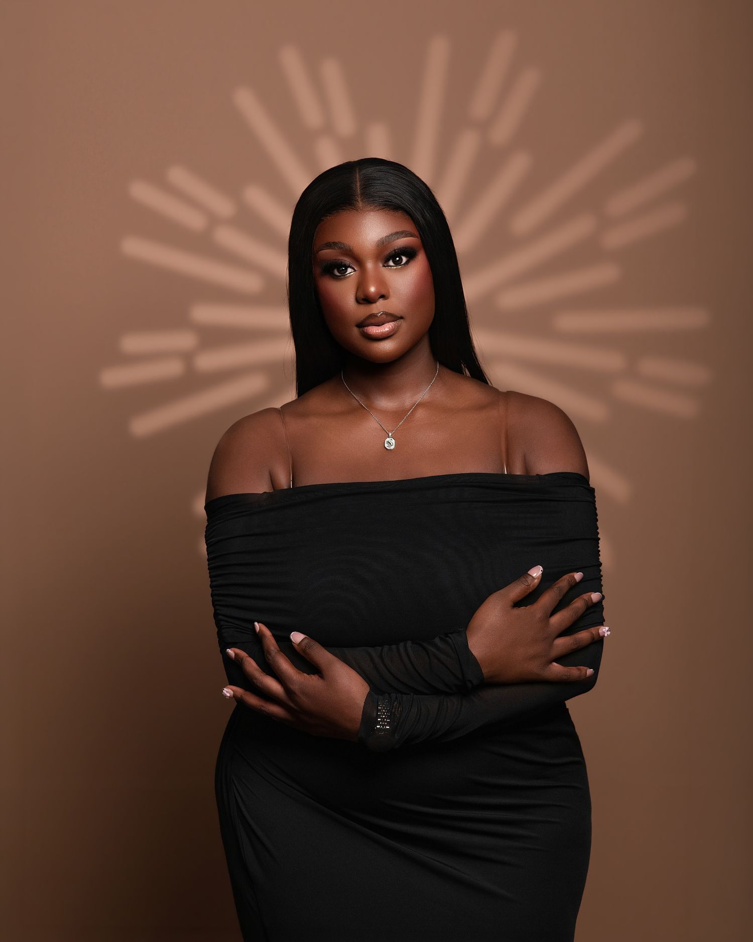 Professional beauty portrait against warm brown background with dramatic lighting and black off-shoulder styling.