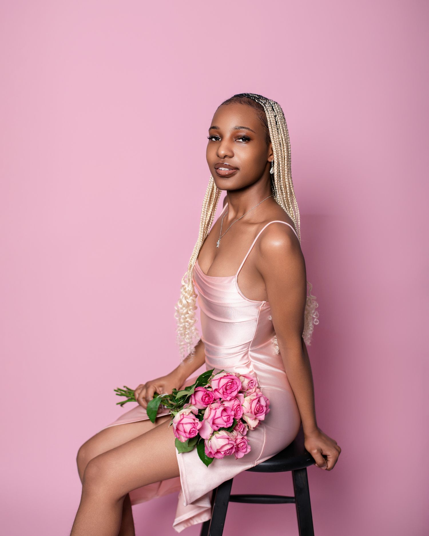 Model in pink silk dress with blonde braids holds pink roses against pink background.