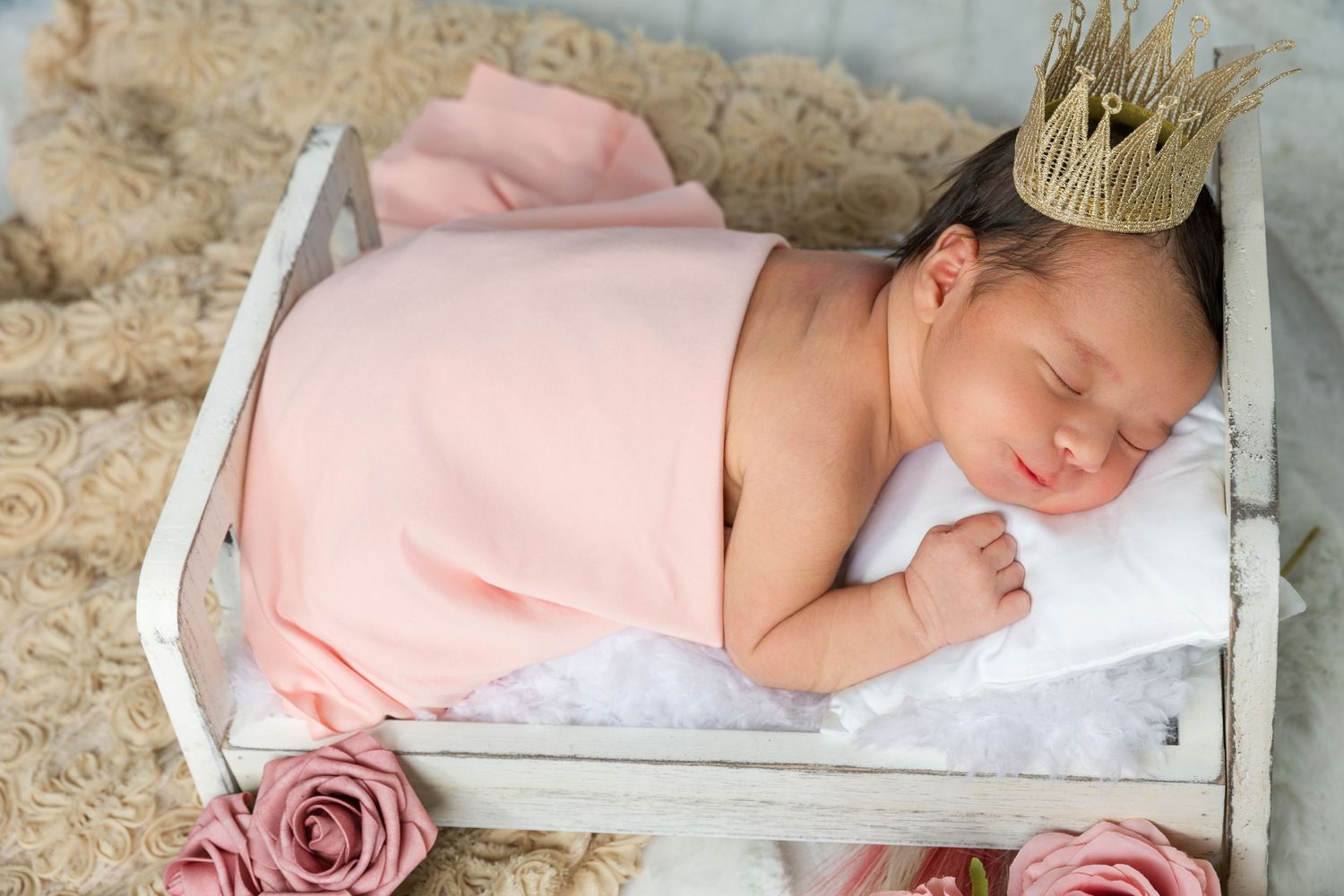 A sleeping newborn wearing a gold crown rests in a rustic white crate with pink blanket and rose decoration.