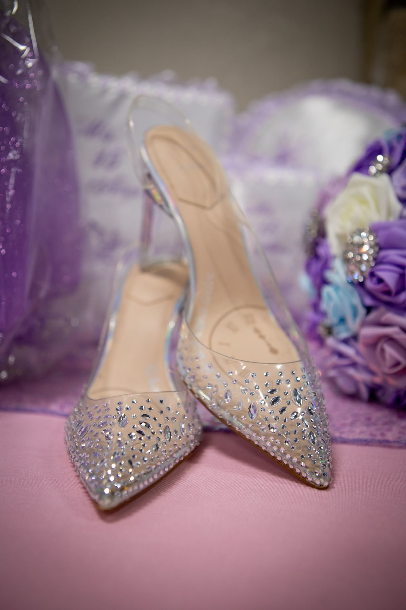 Crystal-embellished pointed toe heels on a pink surface with purple decor in the background.