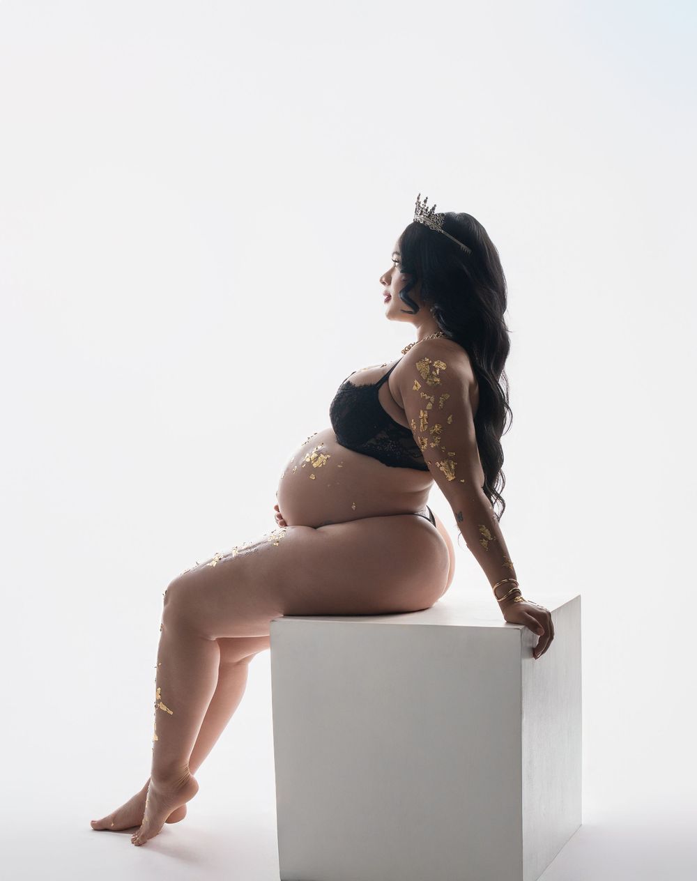 Artistic maternity photo of an expectant mother in black lingerie sitting on a white cube against light background.