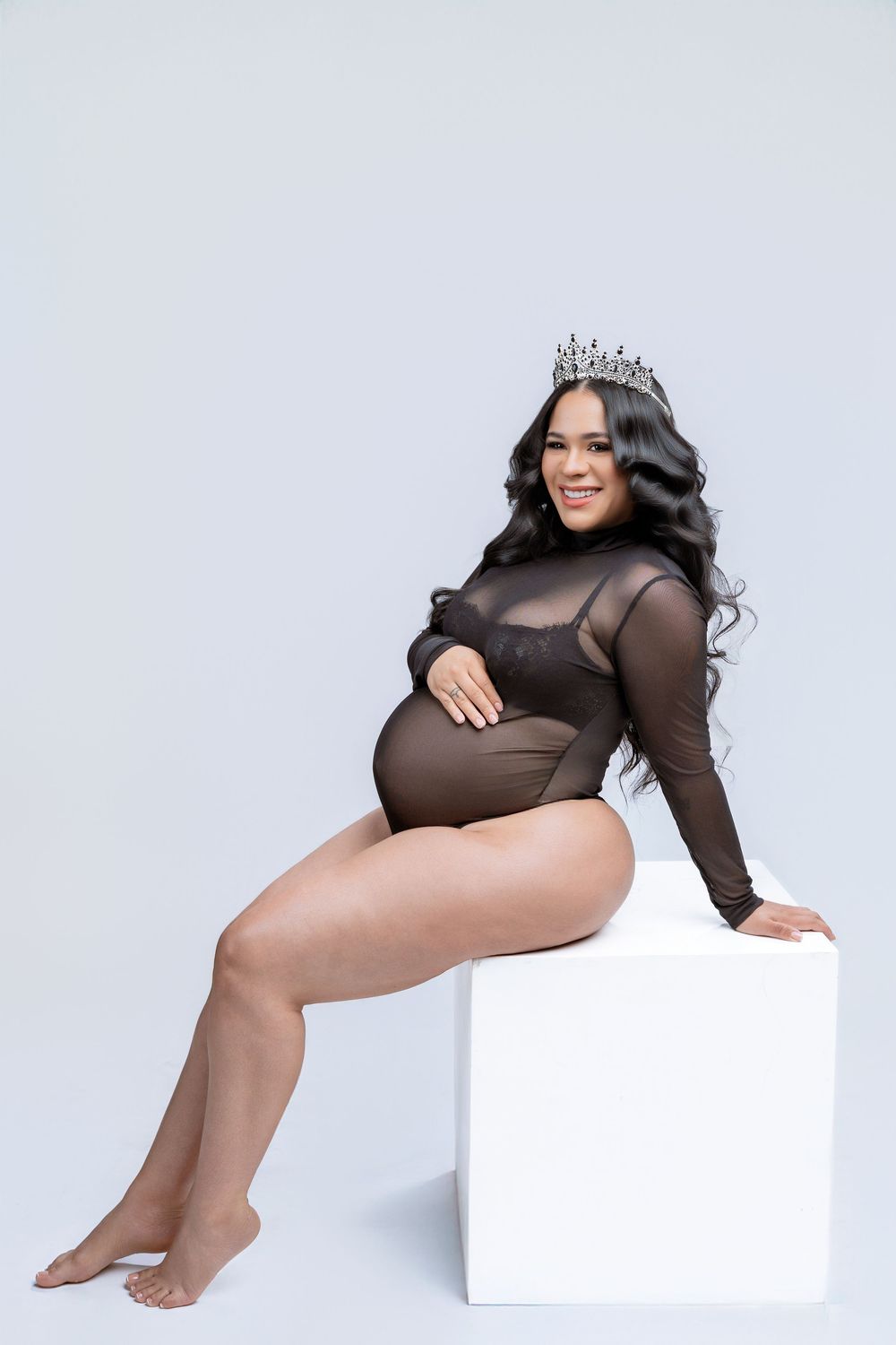 Maternity photo of an expectant mother in a sheer black bodysuit and tiara posing on a white cube against light background.