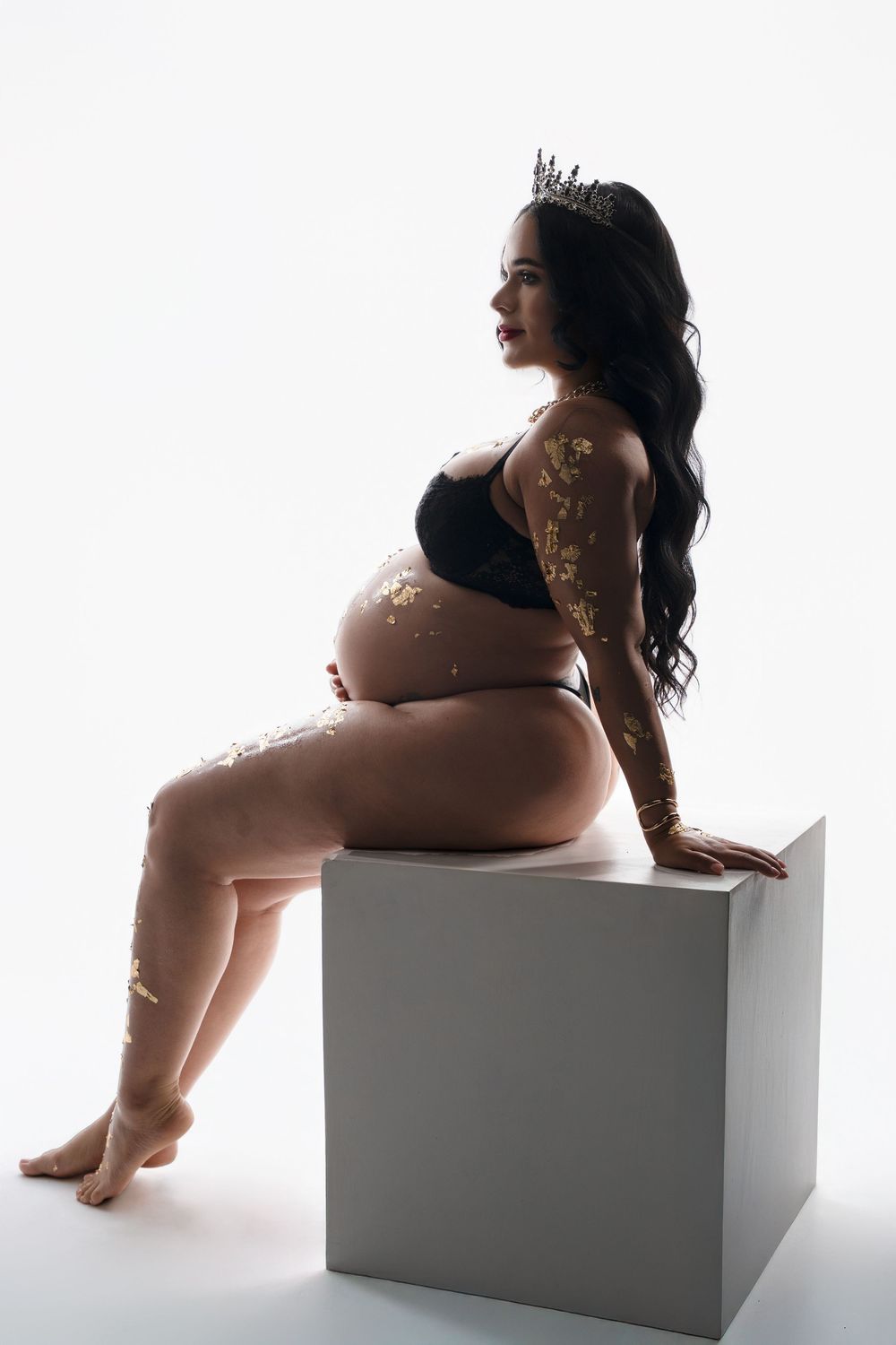 Silhouetted maternity photo of an expectant mother in black lingerie sitting on a white cube against bright background.