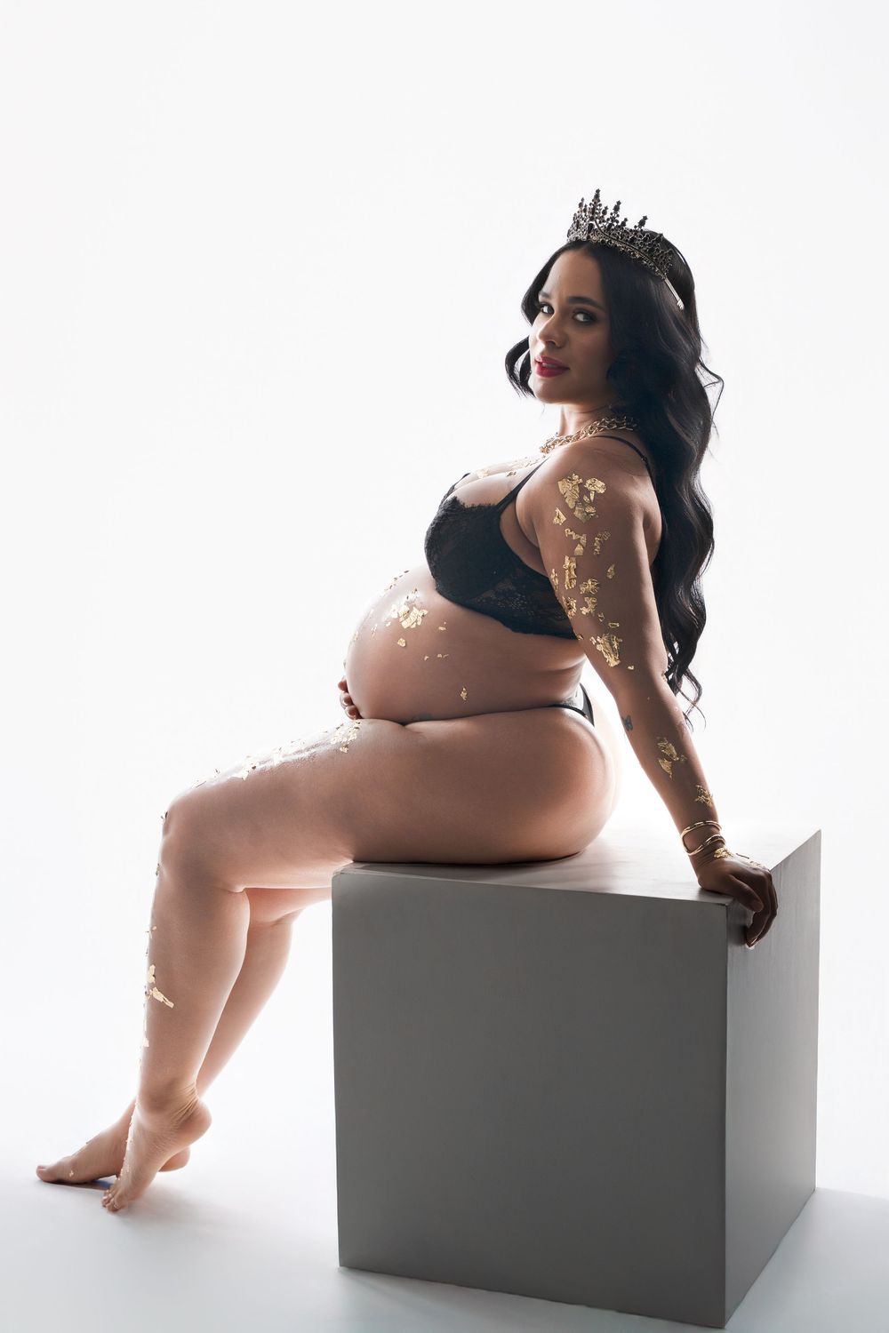 Artistic maternity portrait showing an expectant mother in black lingerie with tattoos posing on a gray cube.
