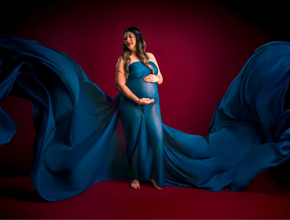 Artistic maternity portrait with flowing blue fabric against dark red studio background.