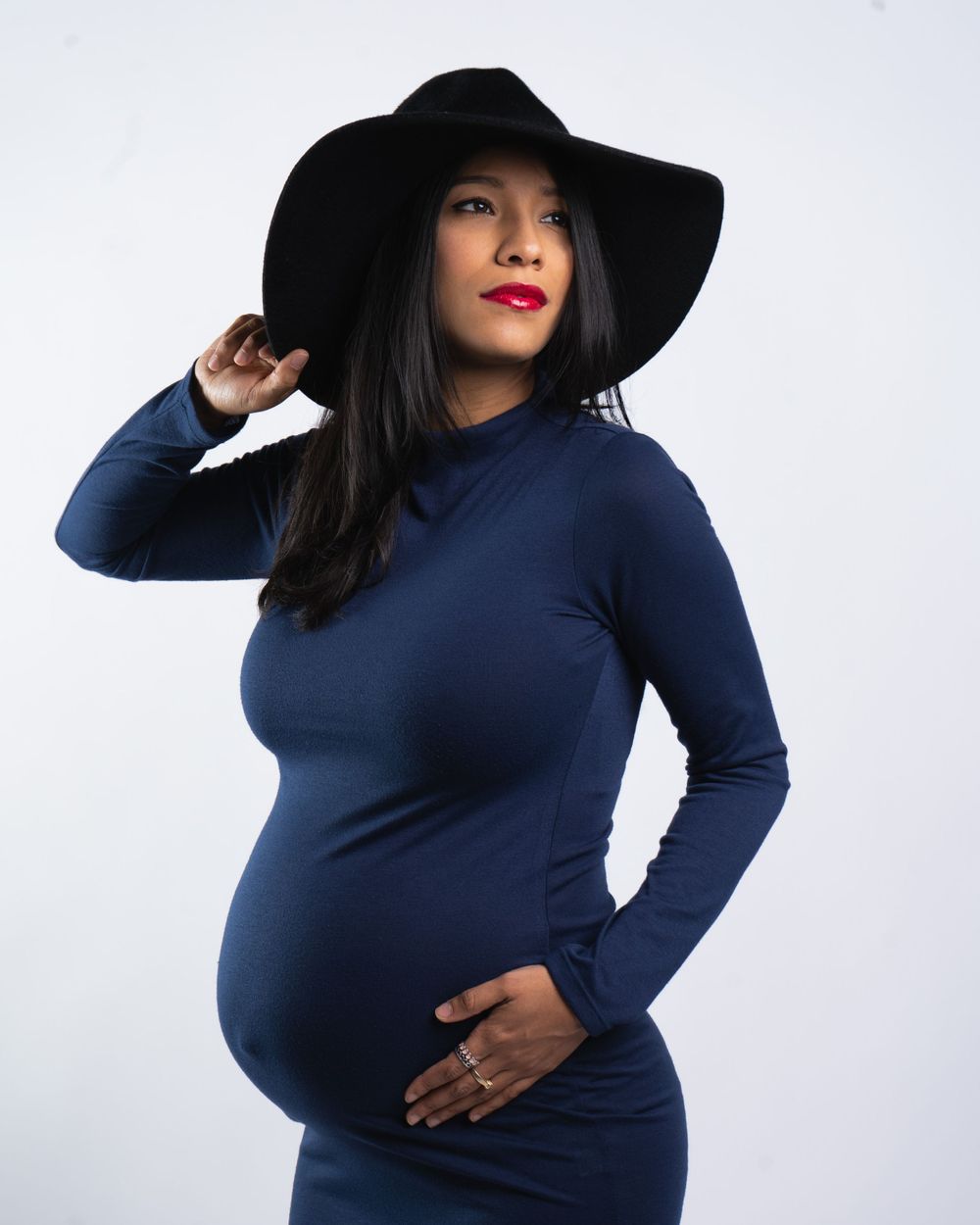 Stylish navy dress maternity portrait with wide-brimmed black hat against white backdrop.