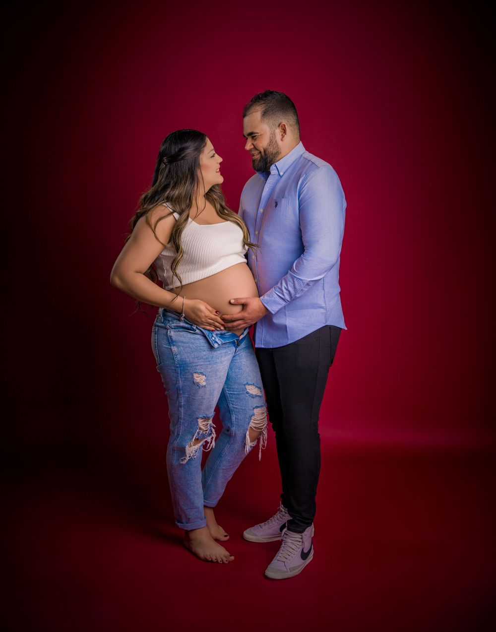 Intimate maternity studio portrait against deep red background with couple in casual attire.