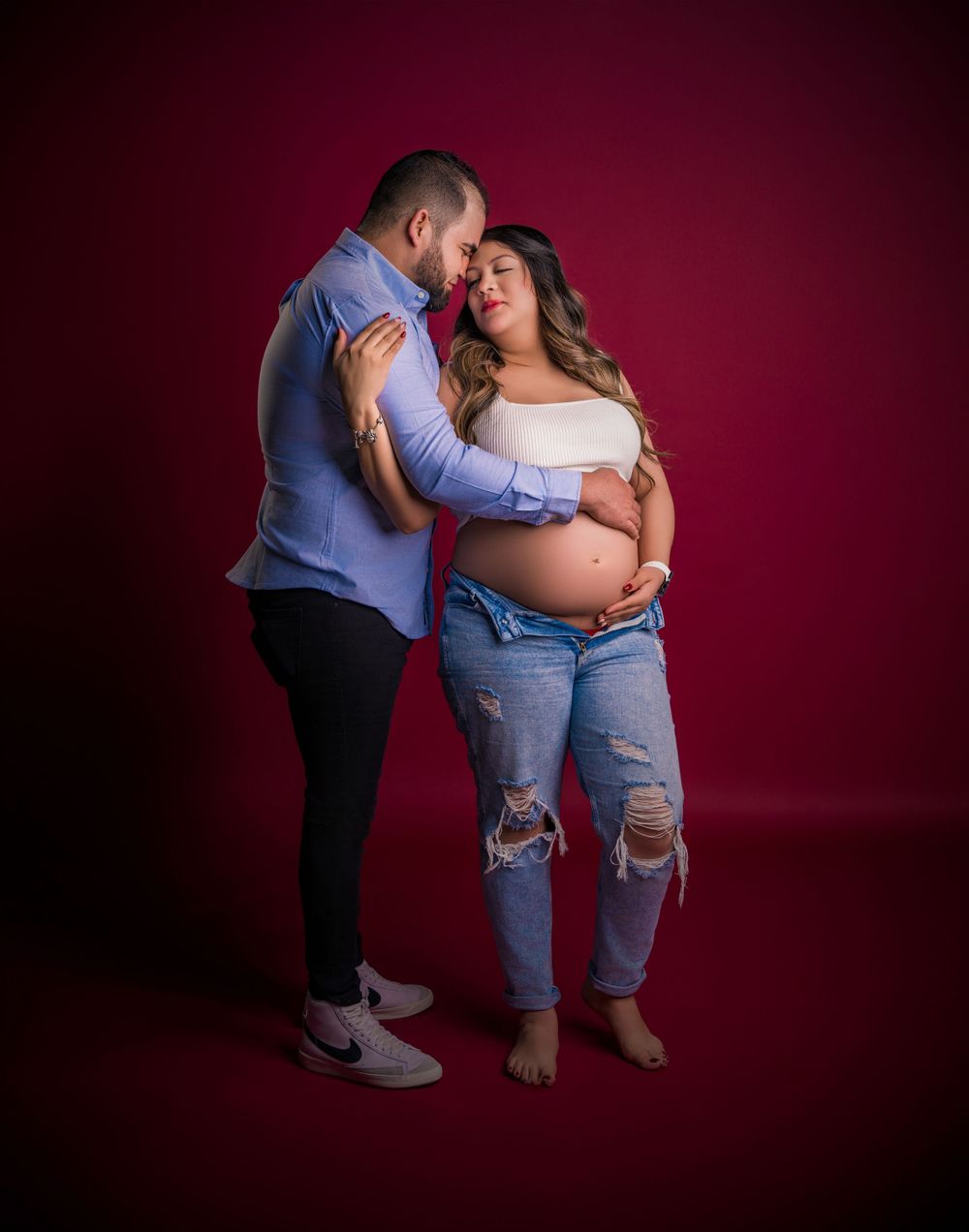 Tender maternity moment captured against burgundy studio backdrop.