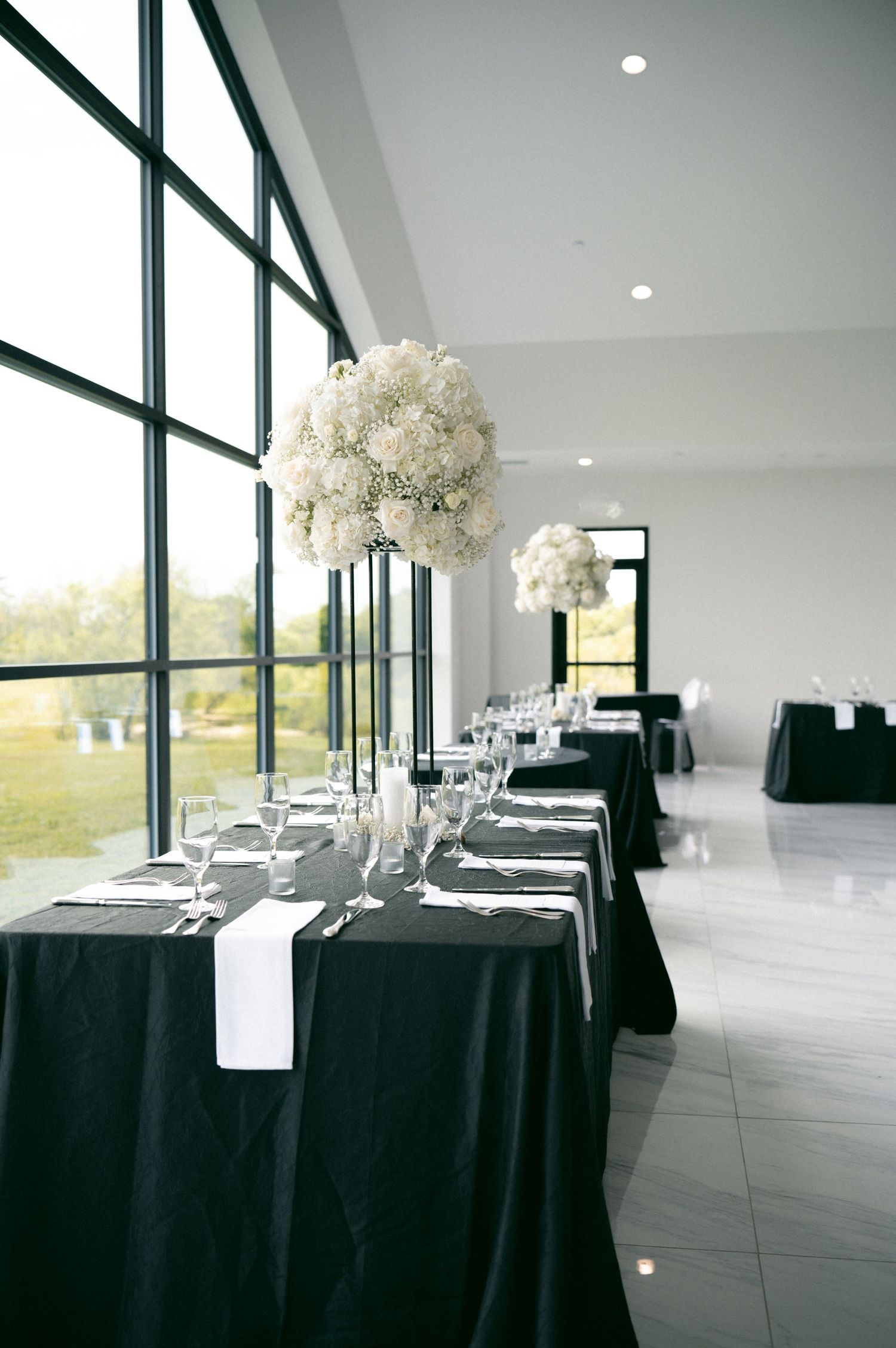 Elegant wedding reception table with black linens, white runners and tall white floral centerpieces beside large windows.