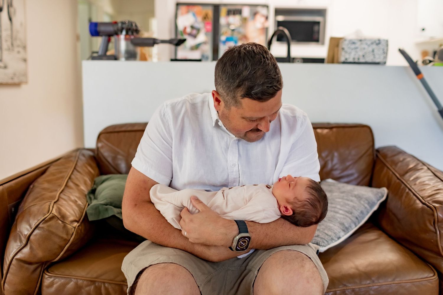 Parent cradles sleeping newborn baby while sitting on brown leather couch.