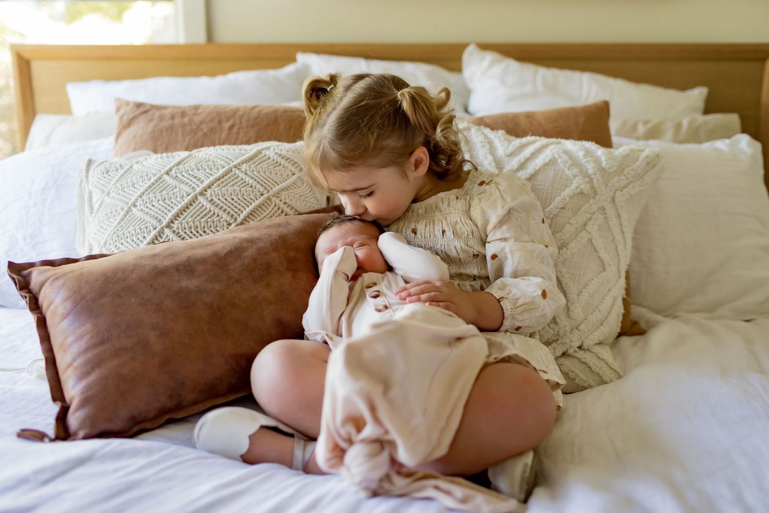 Child with pigtails gently kisses baby while sitting on white bedding.