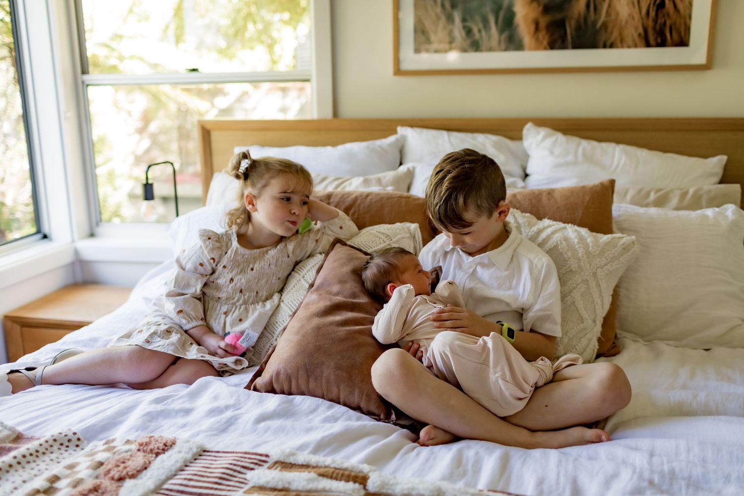 Two children in white clothing sit together on a bed with brown pillows.