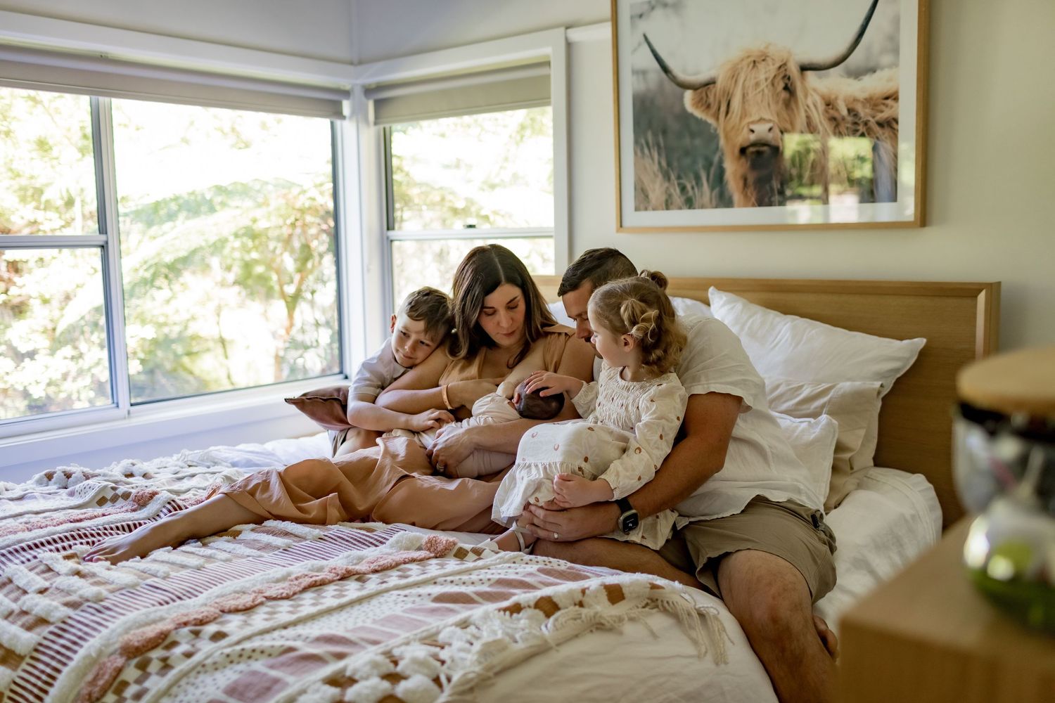 Family relaxing together on bed in bright sunlit bedroom with artwork on wall.