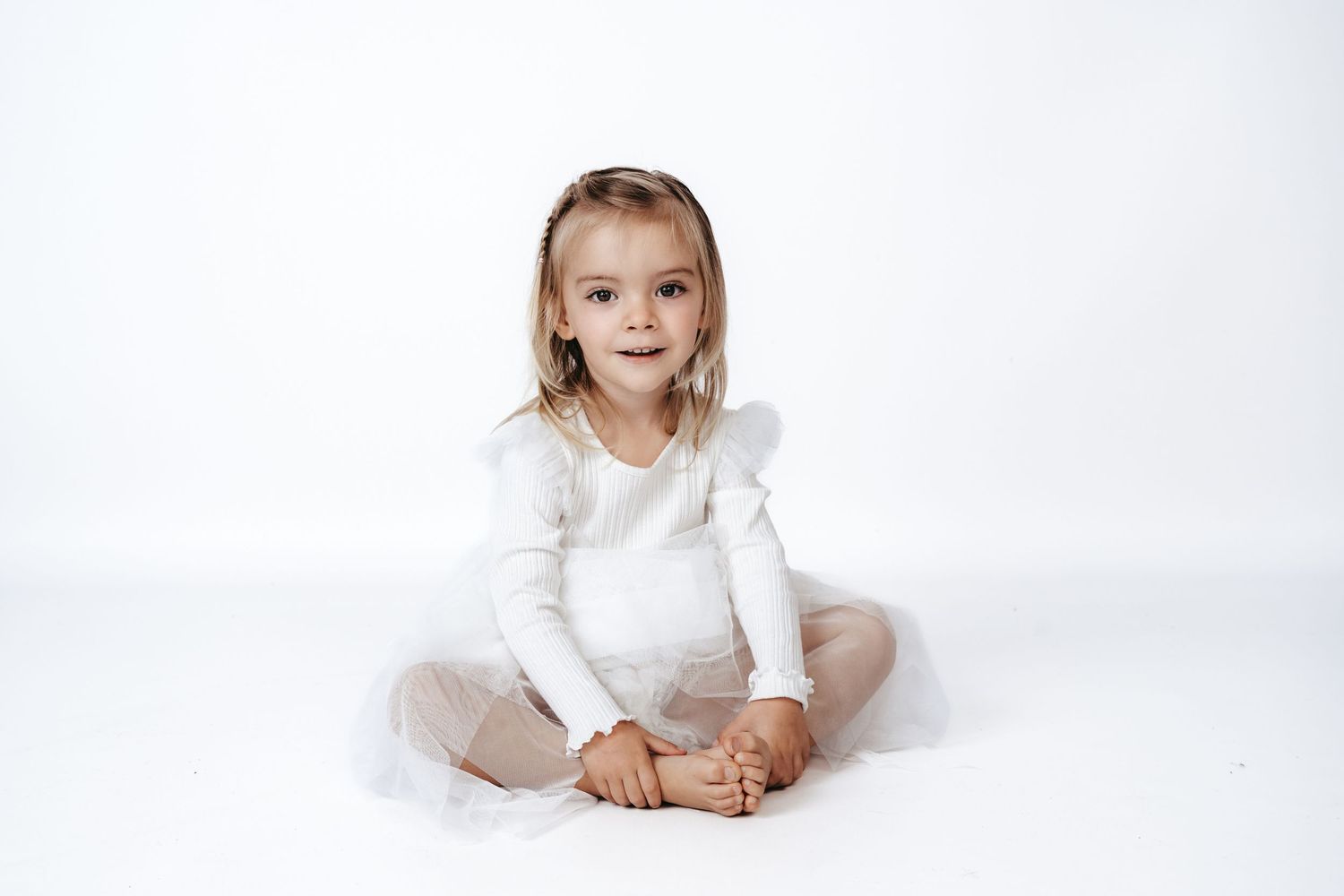 A child in white sits cross-legged on a white floor during a studio photoshoot.