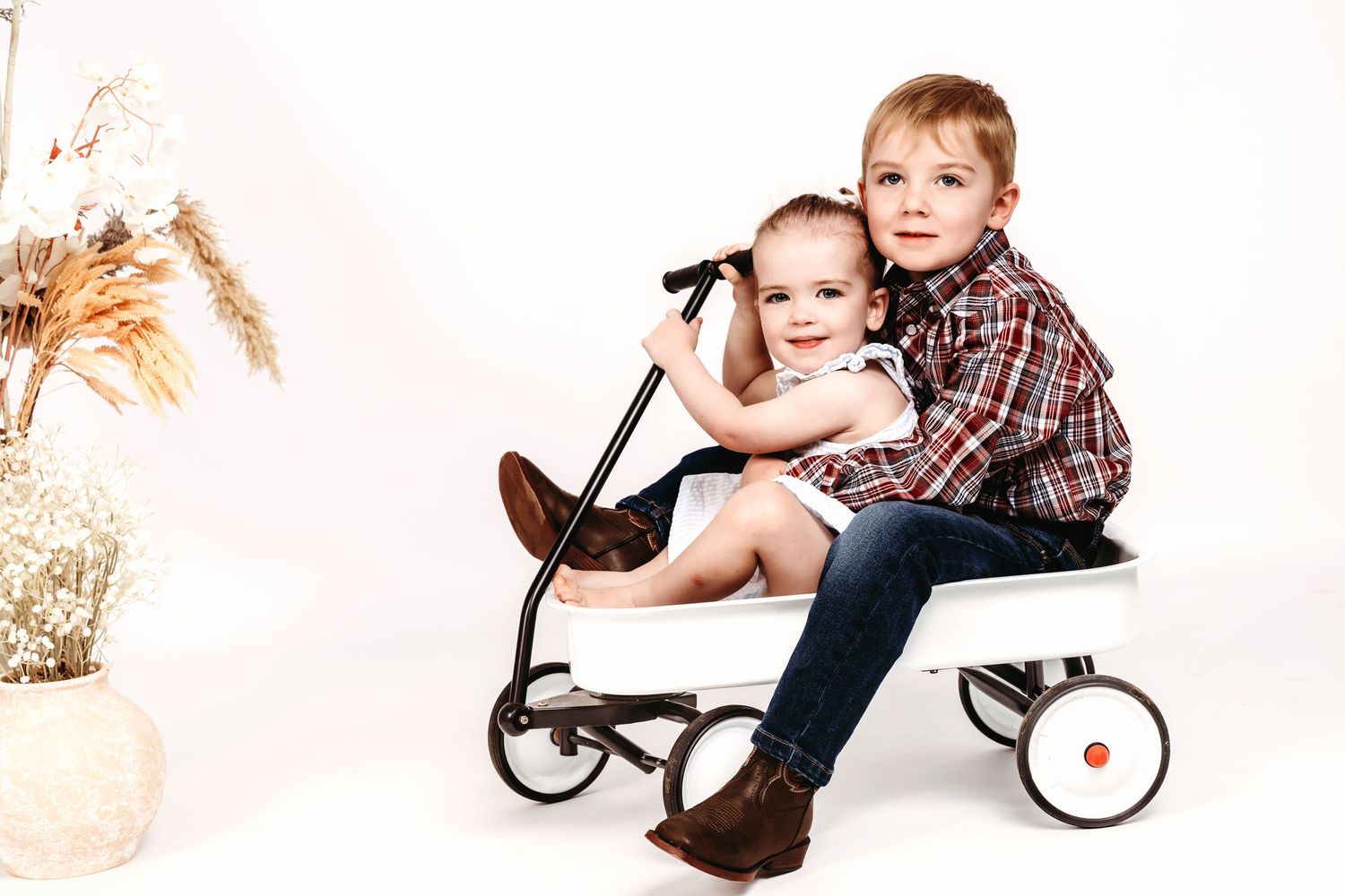 Two children sitting together on vintage red wagon with dried flowers.
