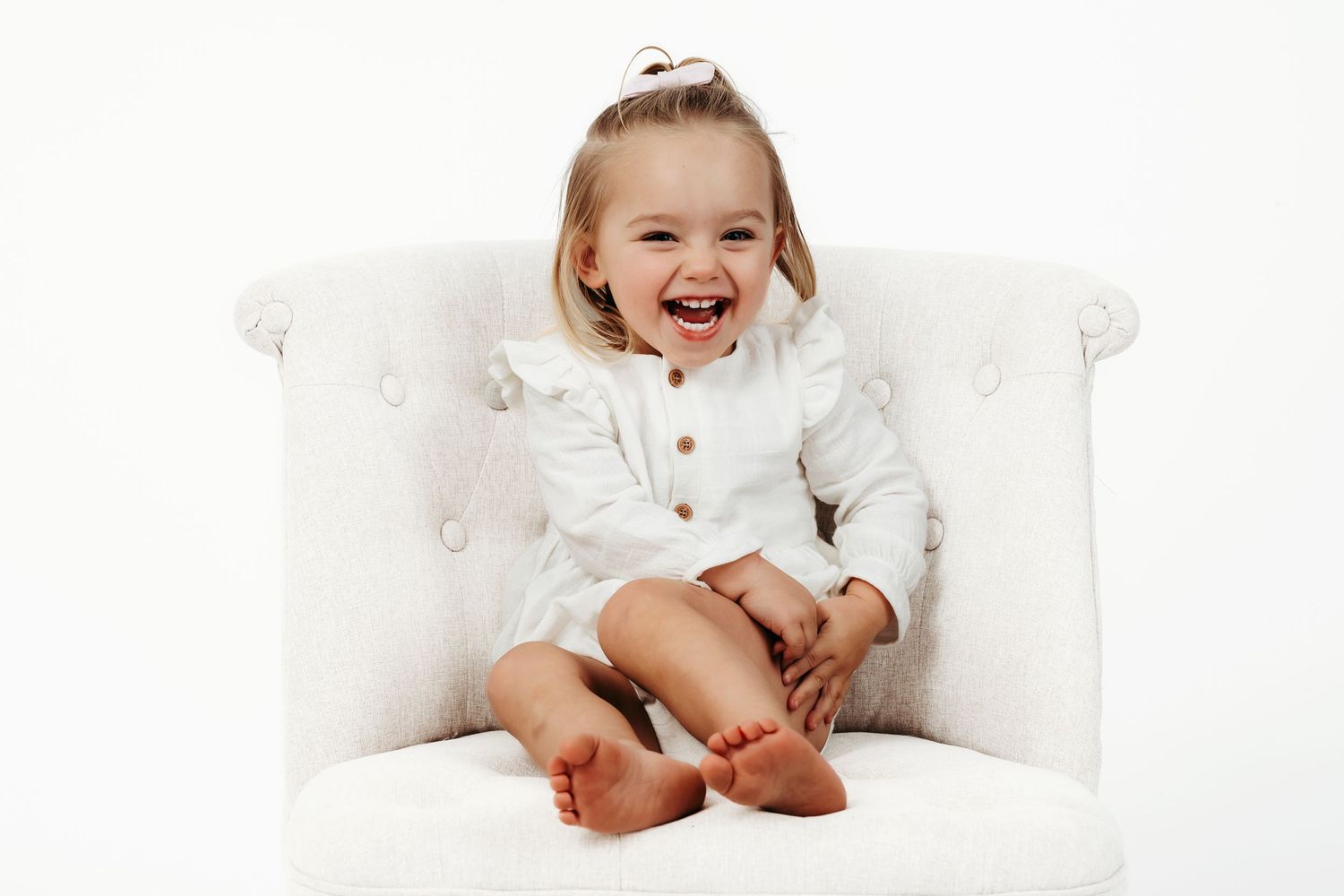 Joyful child in white outfit laughing while sitting on white chair.