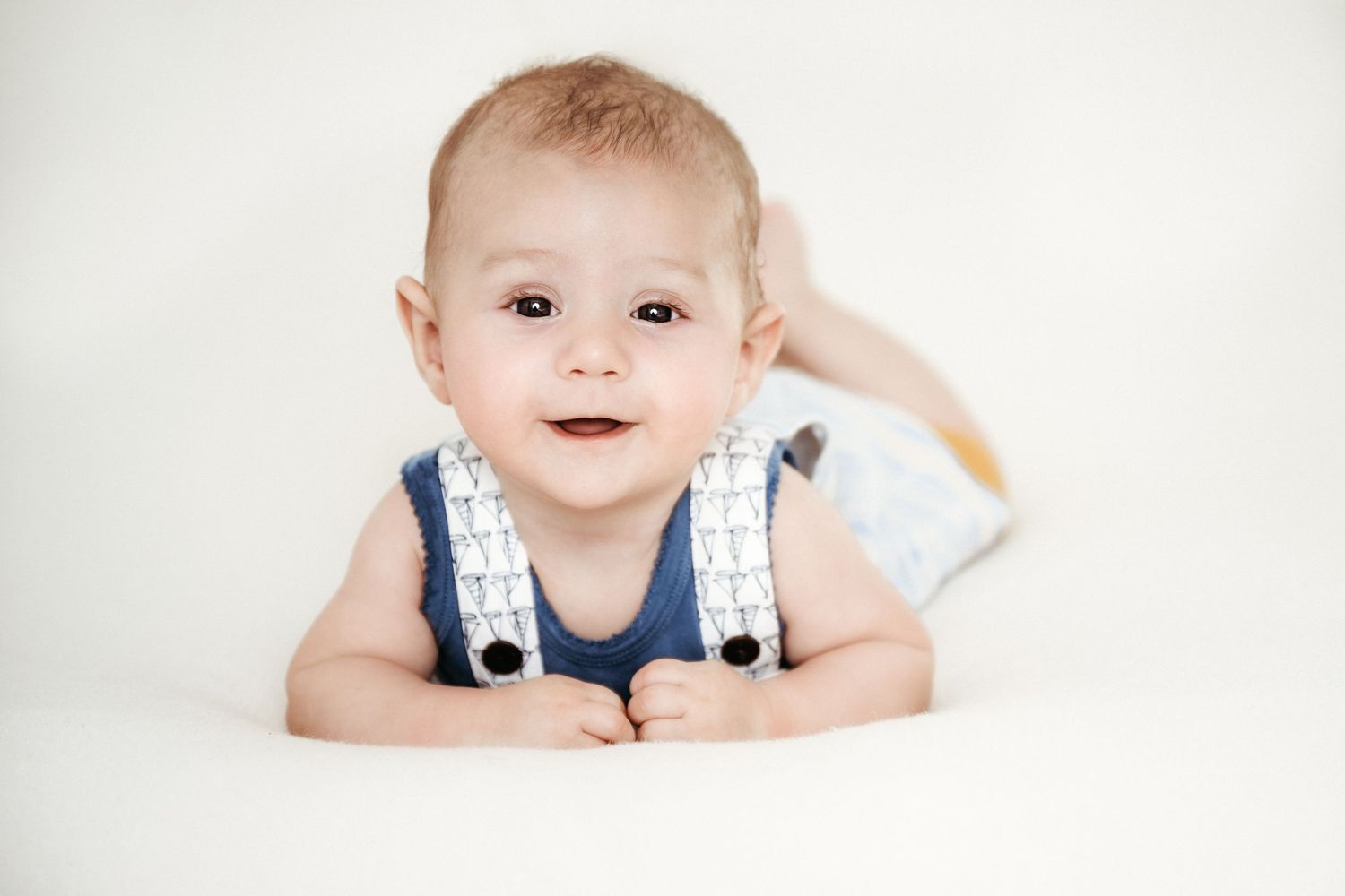 Baby in blue and white overalls lying on stomach against white background.