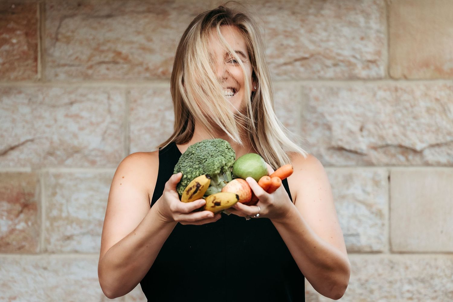 A person in a black tank top holds fresh fruits and vegetables while standing against a stone wall.