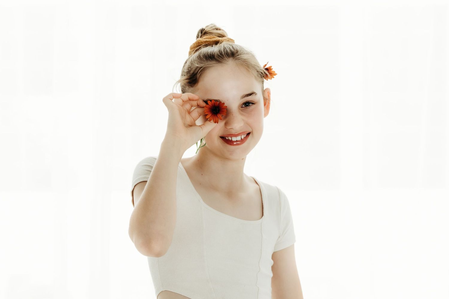 A dancer in a white crop top holds flowers near her face while posing against a bright background.