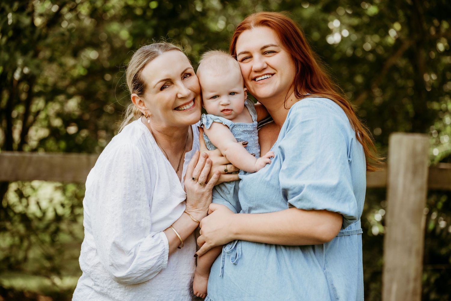 Three people pose for a warm family portrait outdoors in natural lighting against a rustic wooden fence.