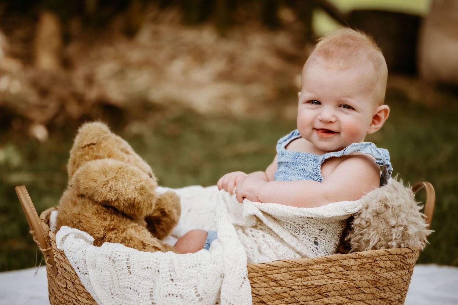 A baby sits in a woven basket with stuffed animals and a white blanket, smiling outdoors.