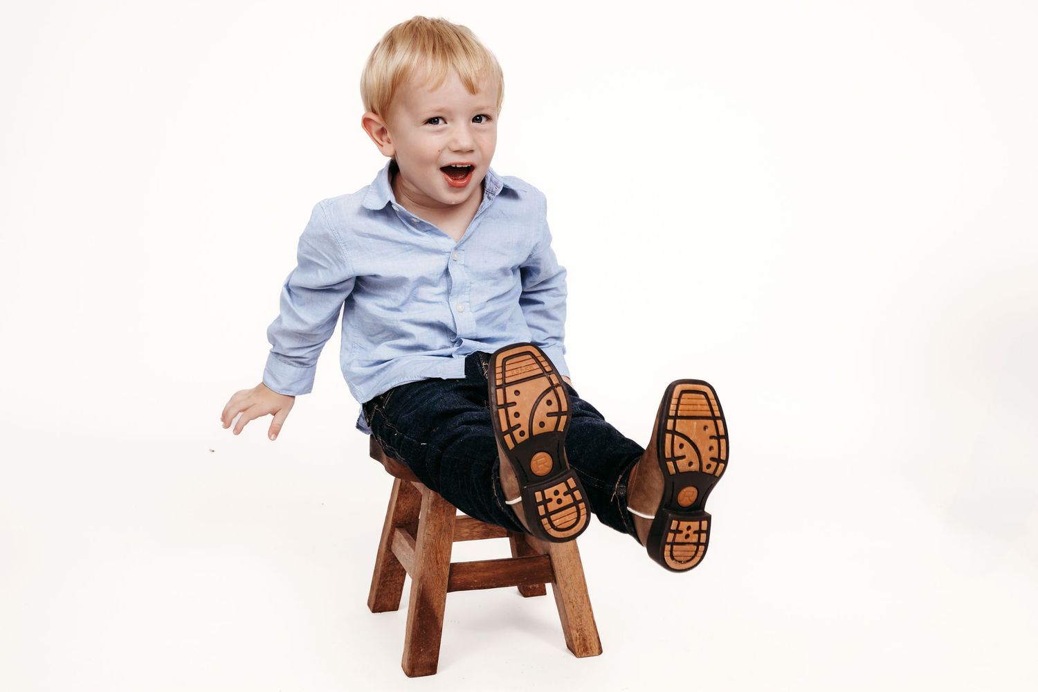 Child in light blue shirt and dark pants sits on wooden stool with feet outstretched against white background.