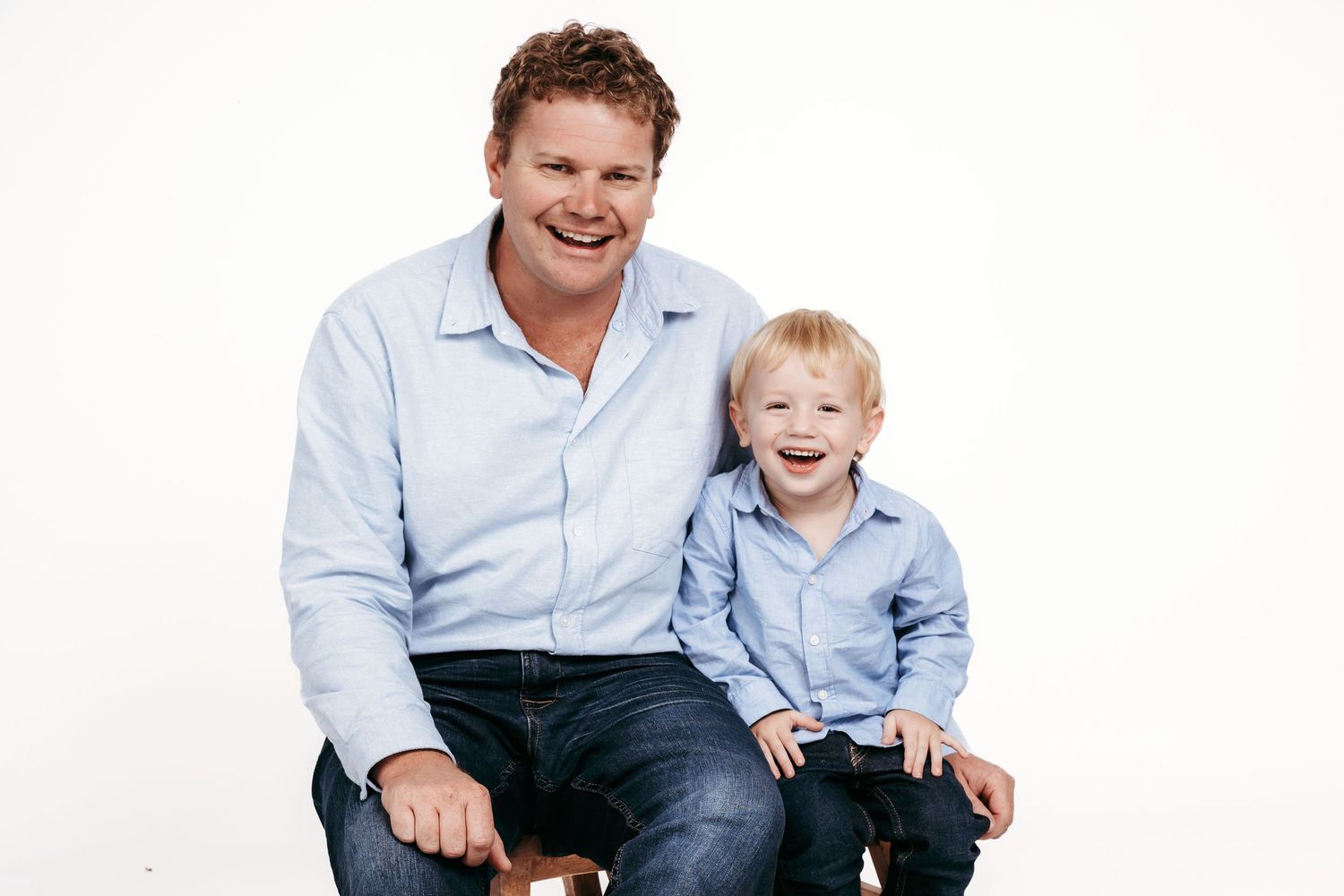 Two people in matching light blue button-down shirts sit together against a white backdrop, sharing a happy moment.