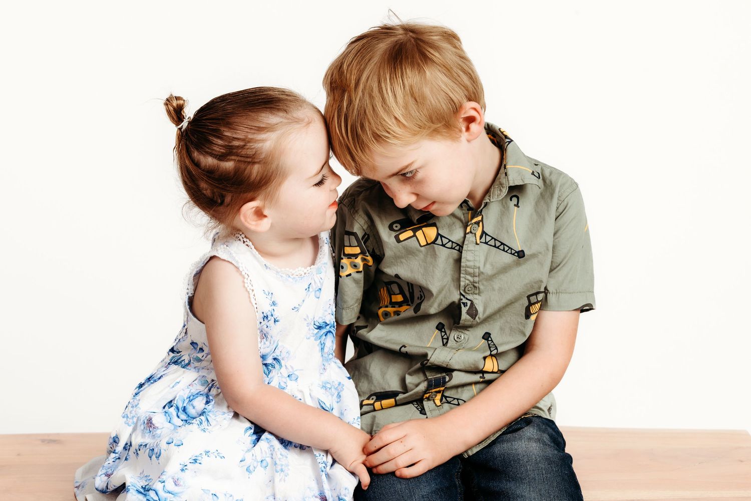 Two children share sweet moment while sitting together against white backdrop.