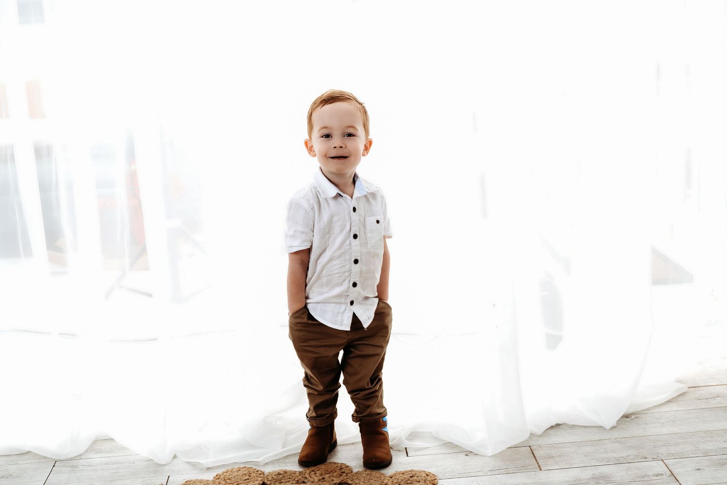 A toddler in a white button-down shirt and brown pants stands against white curtains.