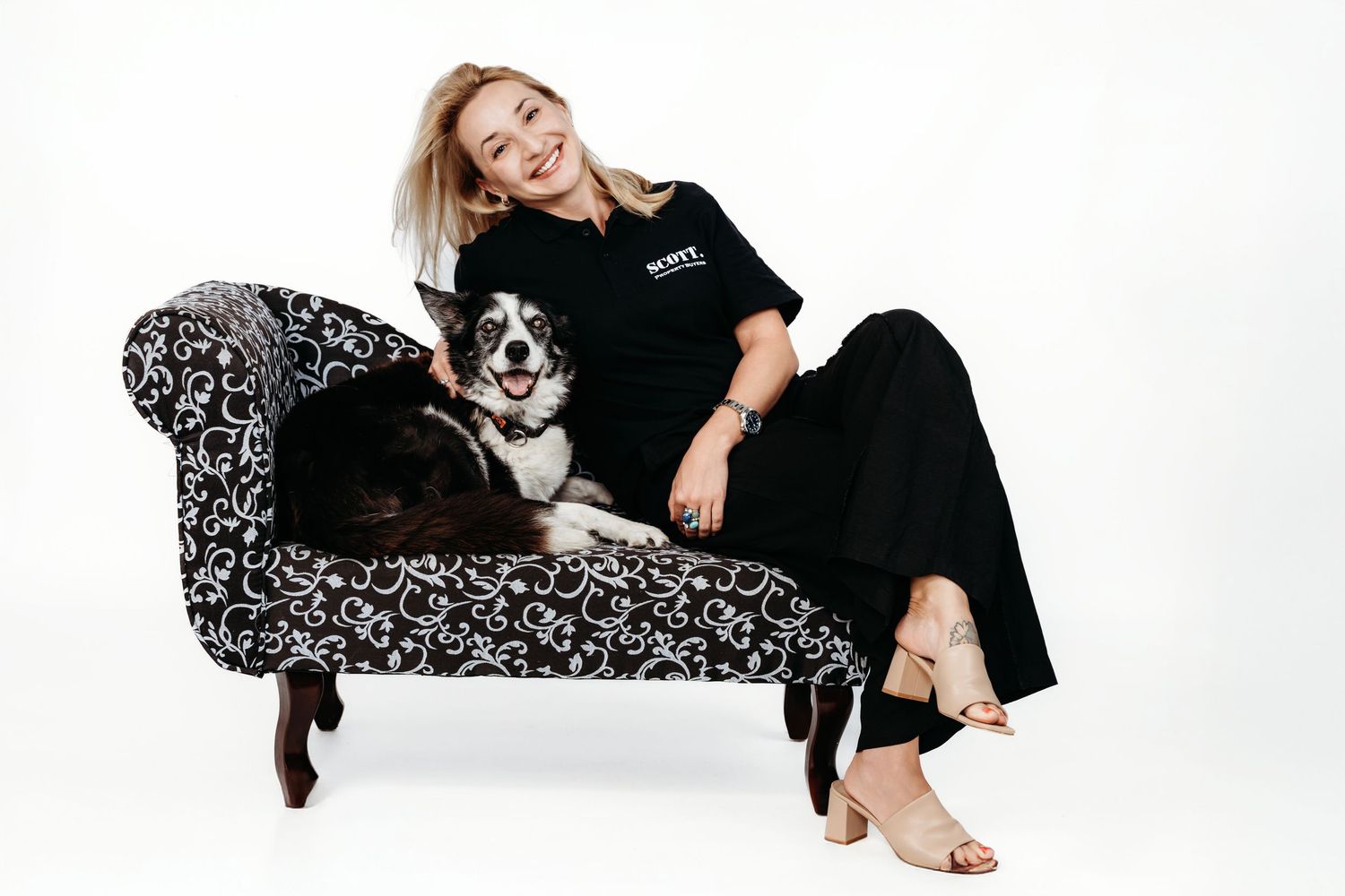 Person lounges on a floral chaise lounge with a black and white Border Collie on white background.