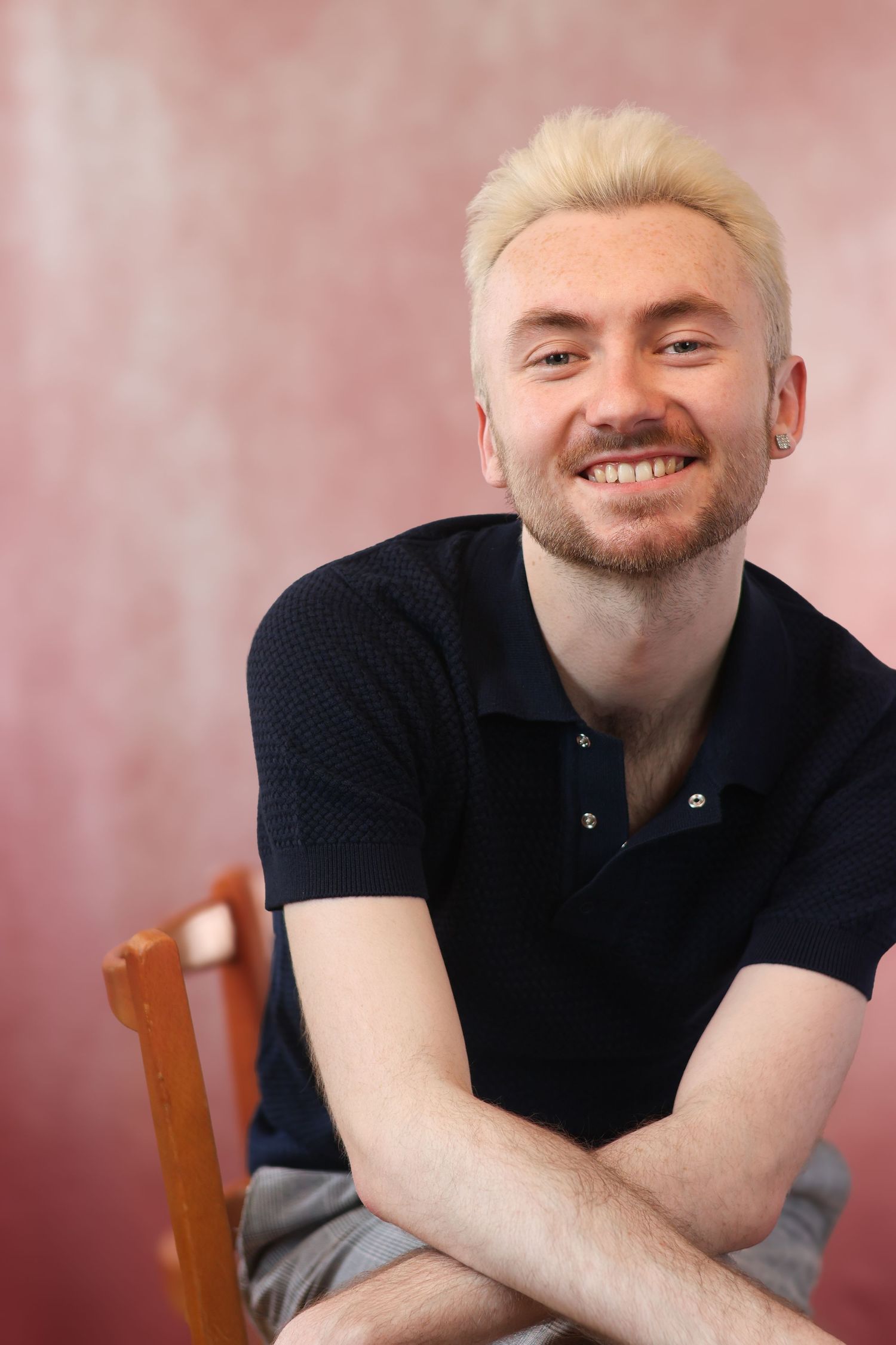 Person with blonde hair in black polo shirt sitting casually on wooden chair.