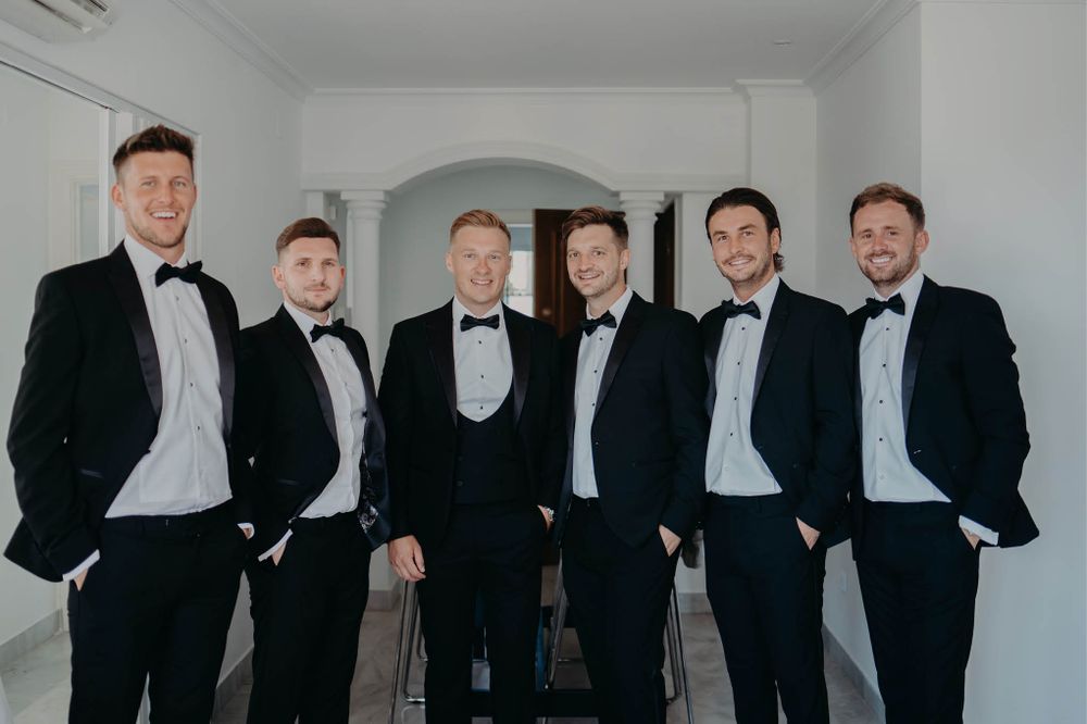Groomsmen in black tuxedos pose together in an elegant white interior.